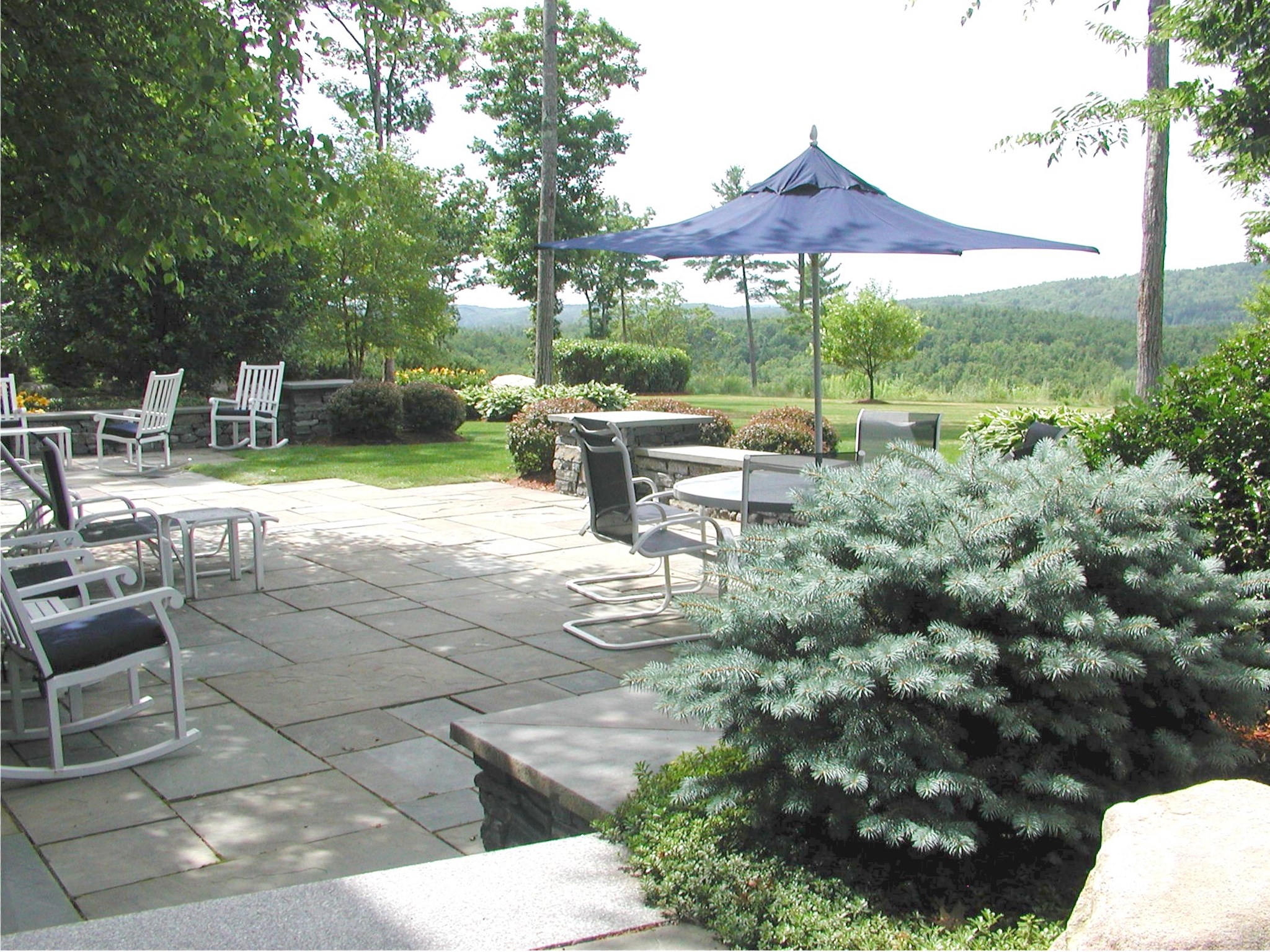 A serene patio with stone flooring, outdoor seating, and a blue umbrella overlooks lush greenery and distant hills under a clear sky.