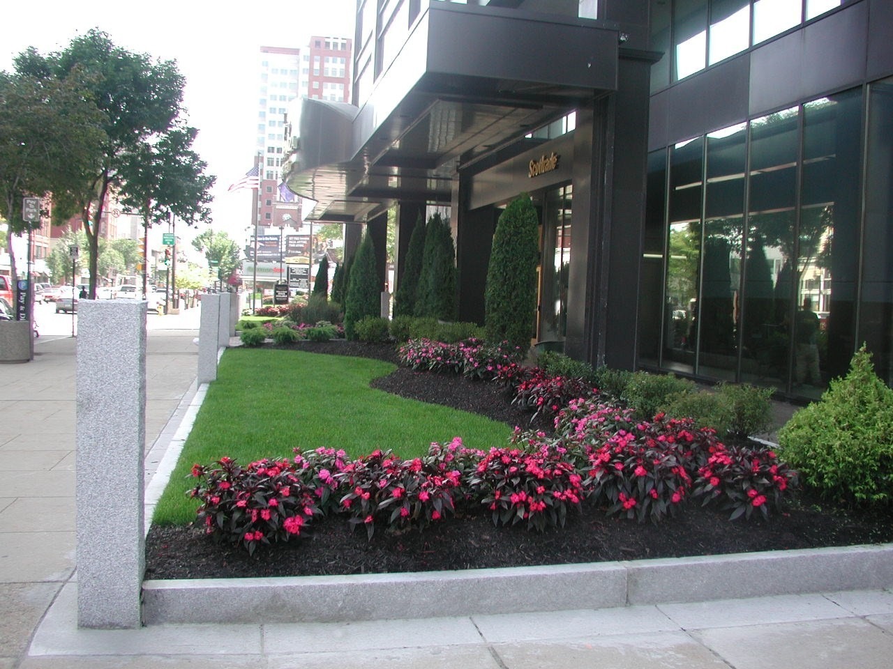 Urban scene with modern glass building, manicured lawn, and vibrant flower bed. Tall structures in the background, street visible with walking person.