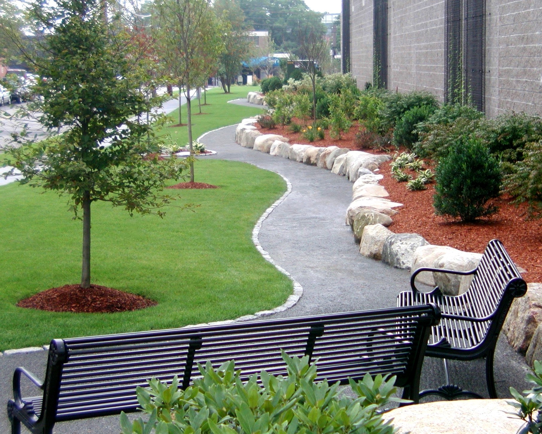 A tranquil park scene with a winding path, benches, landscaped gardens, and strategically placed rocks, creating a serene outdoor environment.