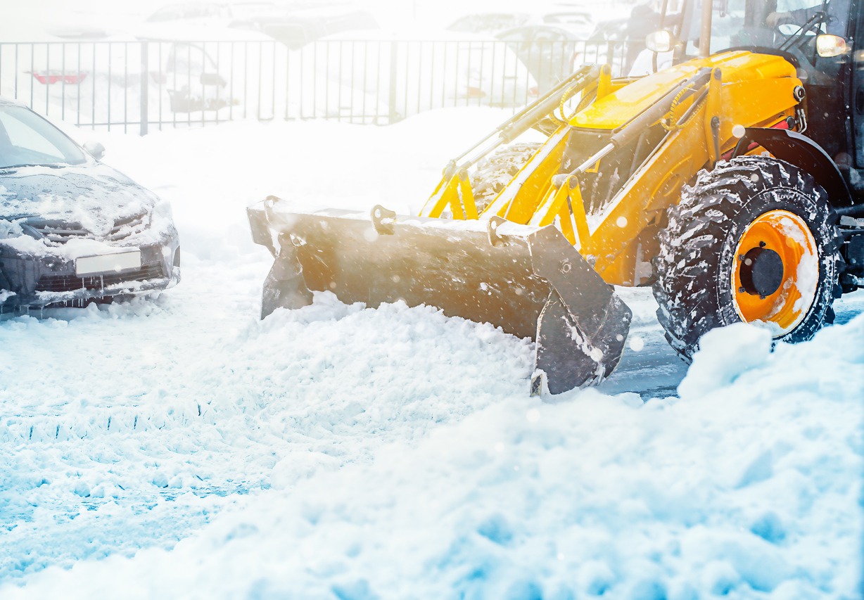 A yellow snowplow clears snow from a parking lot with parked cars nearby. Bright sunlight reflects on the snowy landscape.