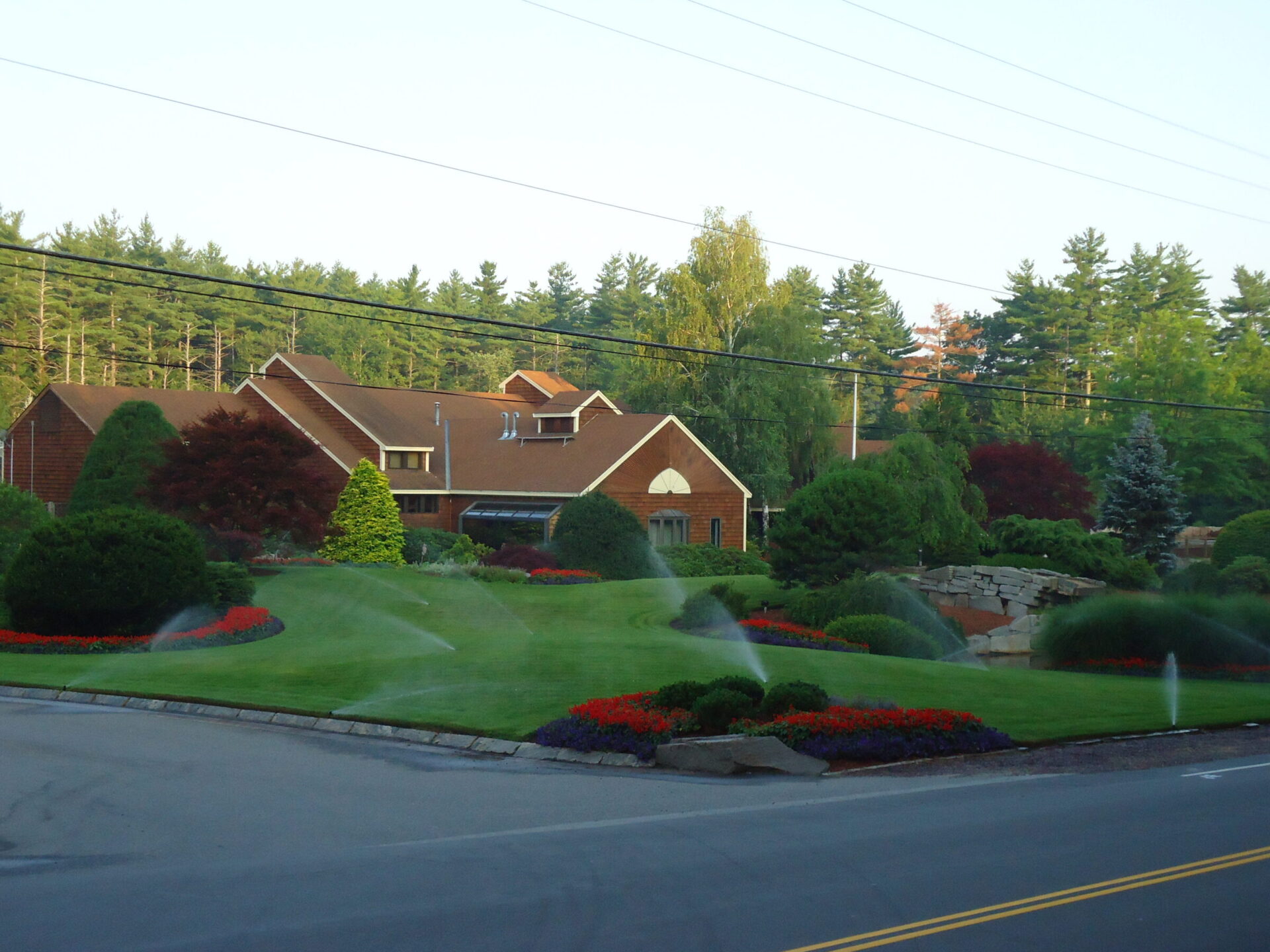 A suburban house with a well-manicured garden, colorful flowers, and sprinklers, surrounded by trees under a clear sky. No landmarks visible.