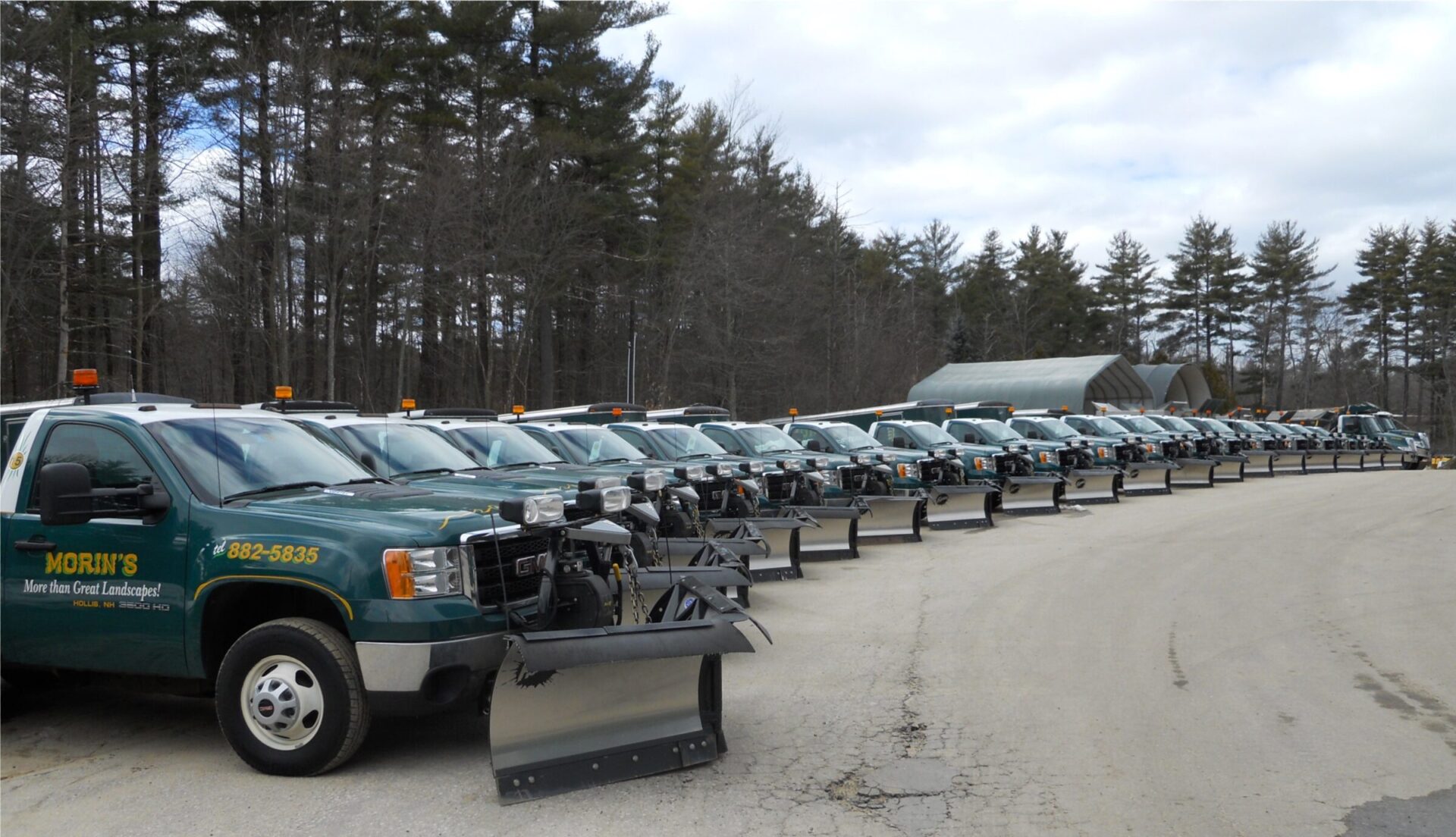 A fleet of snowplow trucks is lined up on a paved road near a forested area under a cloudy sky.