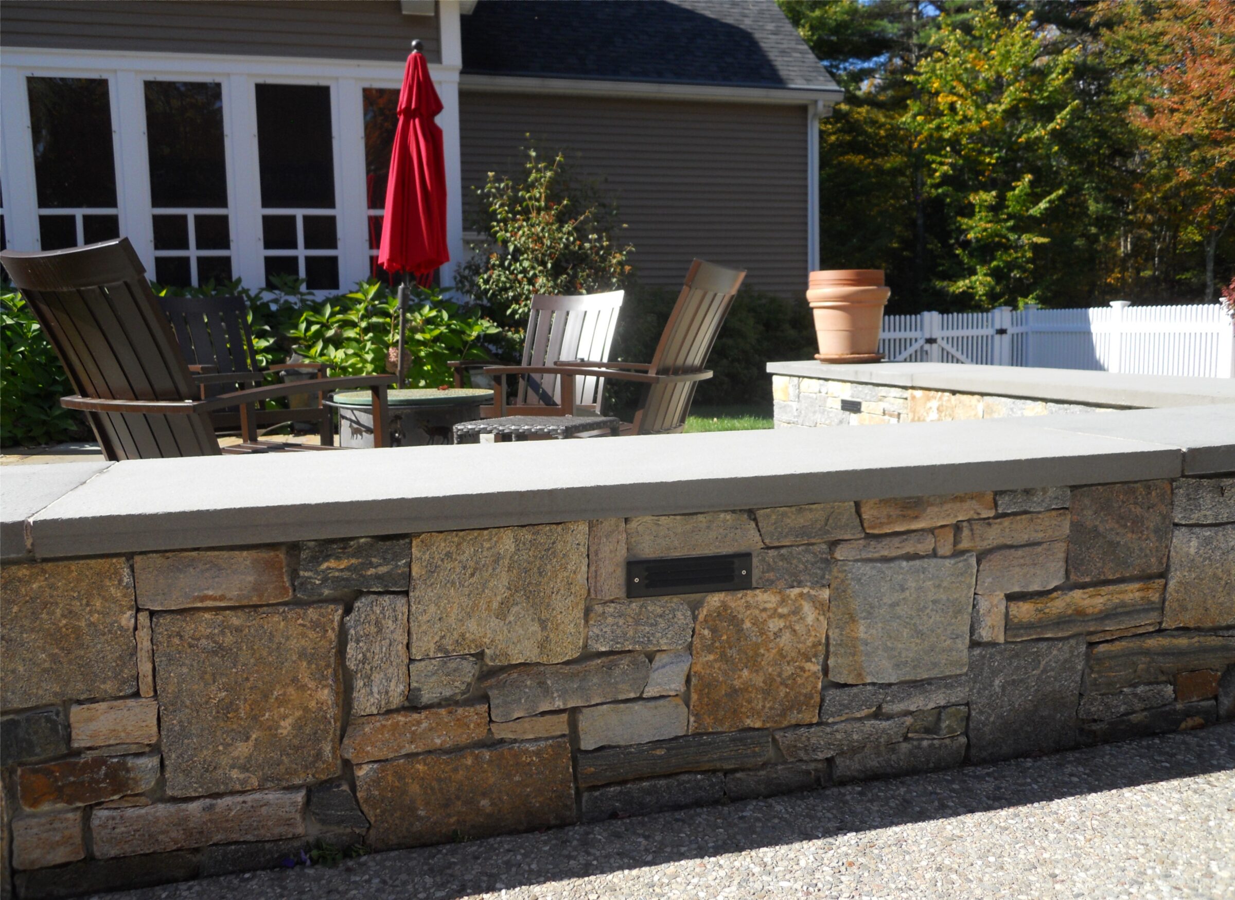 A stone patio features wooden chairs, a red umbrella, and potted plants, set against a house with large windows and greenery.
