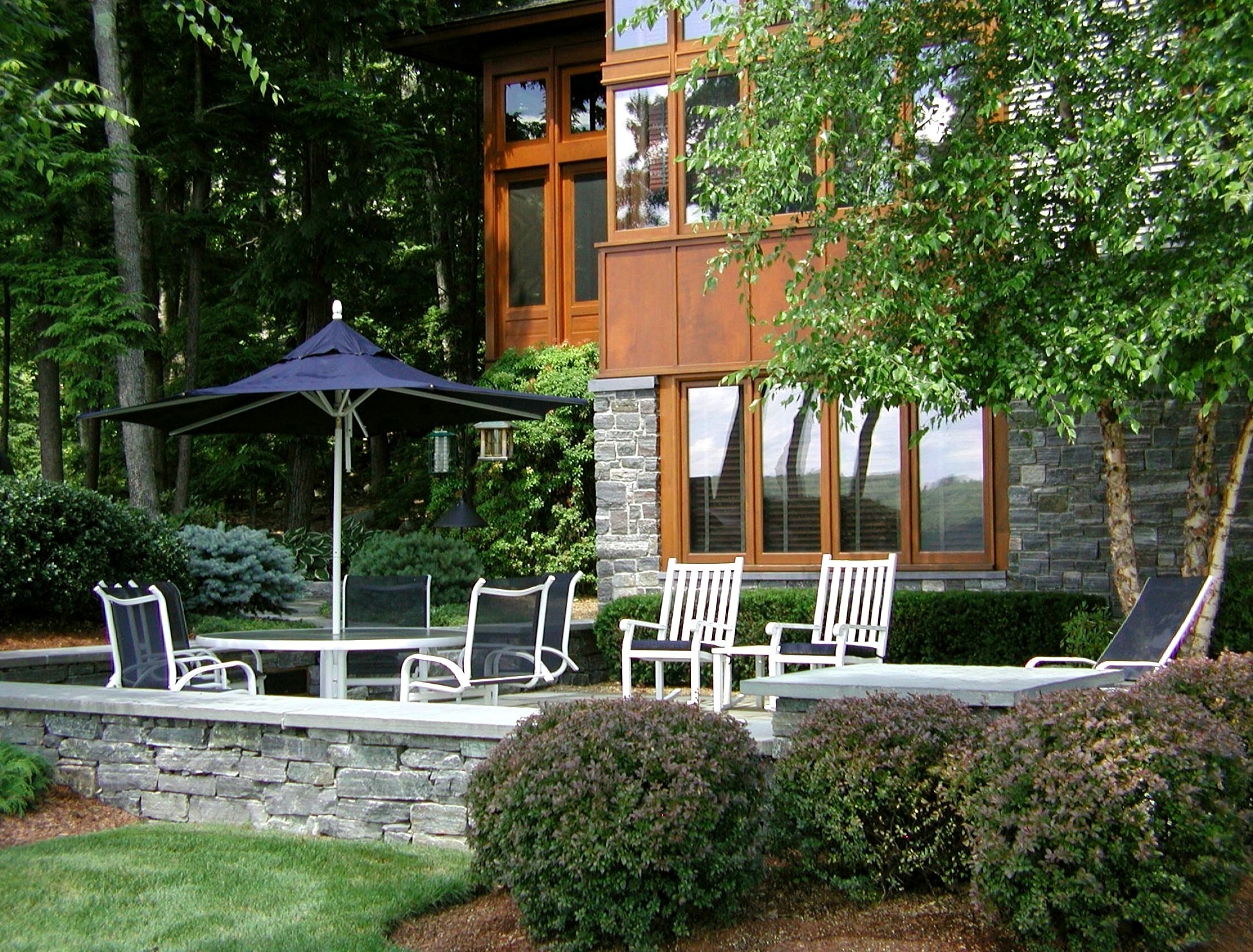 A modern stone patio features a seating area with chairs, a table, and a large umbrella, surrounded by lush greenery and trees.