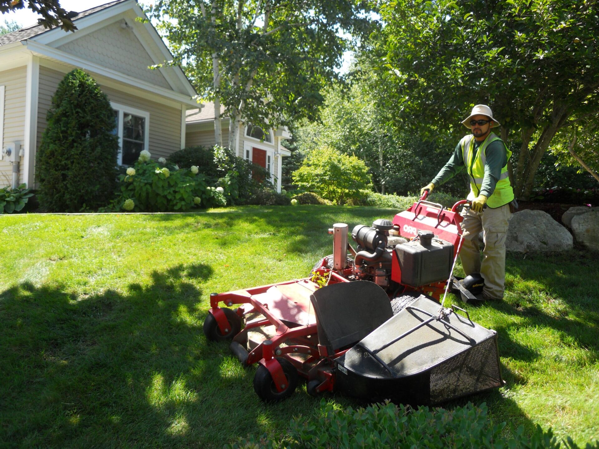 A person operates a red lawnmower on a sunny, grassy slope near a residential house, surrounded by trees and shrubs.