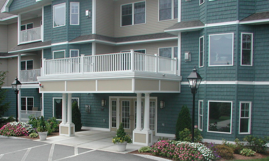 A modern building with green siding, white railing, balcony, and flowering shrubs beside the entrance. Two decorative lamps flank the pathway.