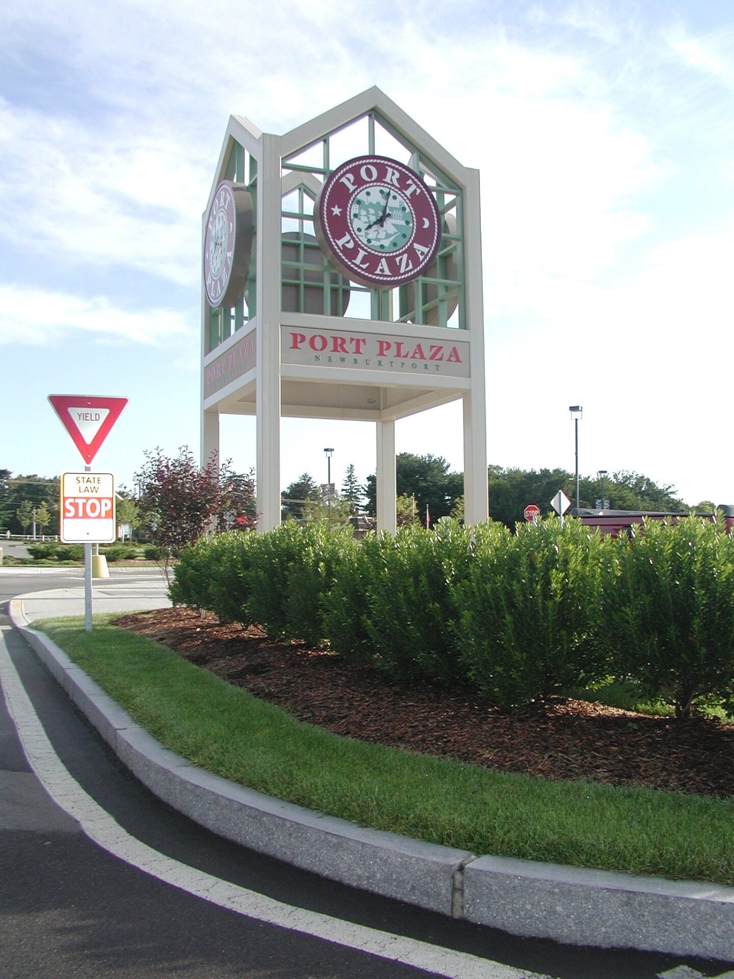 Sign for Port Plaza Shopping Center stands near a yield sign. Well-maintained shrubbery and clear blue sky in the background.