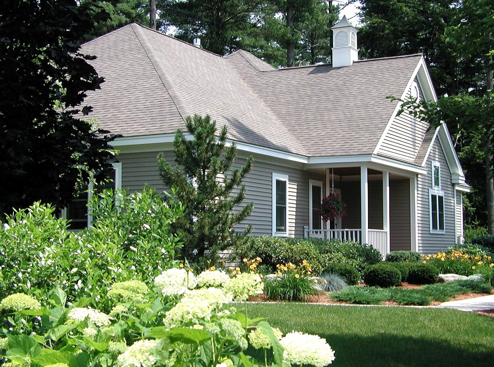 A cozy house with a gable roof, surrounded by lush greenery and blooming flowers, nestled in a wooded area. No landmarks present.
