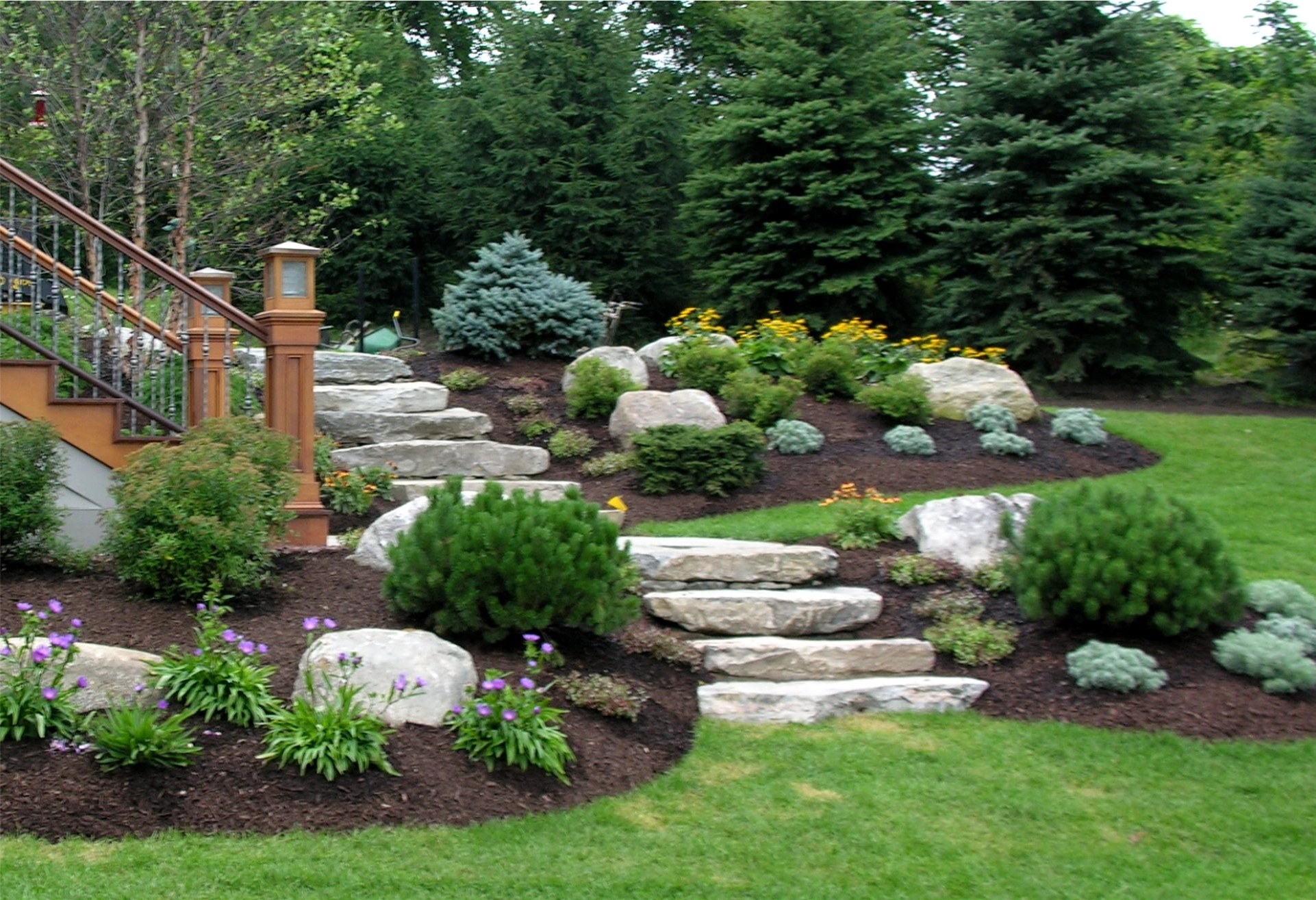 Stone steps lead up a landscaped garden with flowers, shrubs, and trees. Wooden railing visible on the left side.