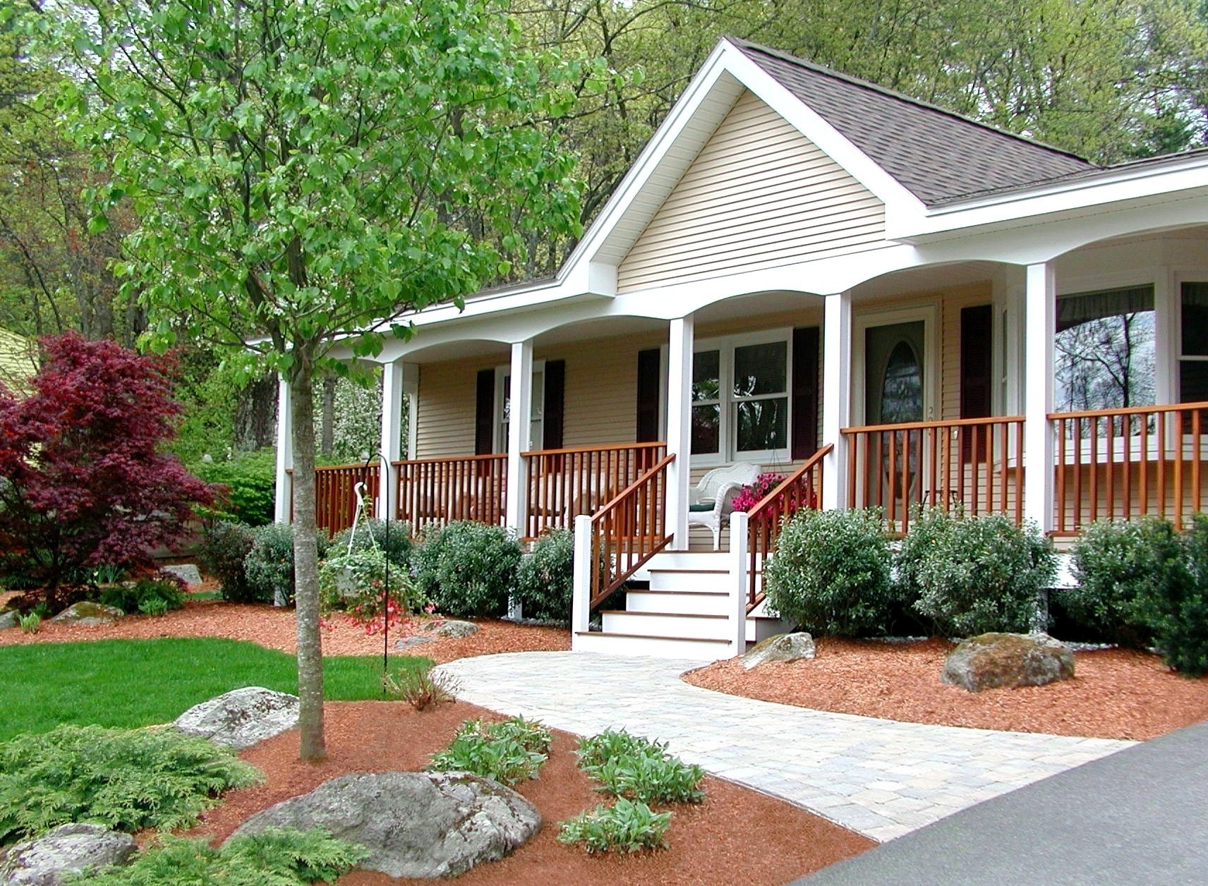 A cozy suburban house with a porch, surrounded by lush greenery and landscaped gardens, features a welcoming path leading to the entrance.