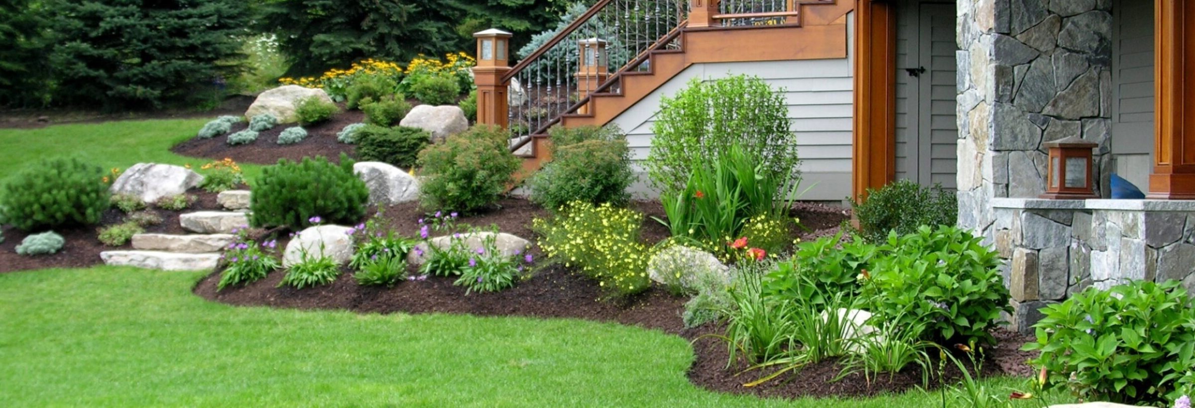 A beautifully landscaped garden with stone steps, colorful flowers, shrubs, and a wooden porch attached to a stone exterior house.