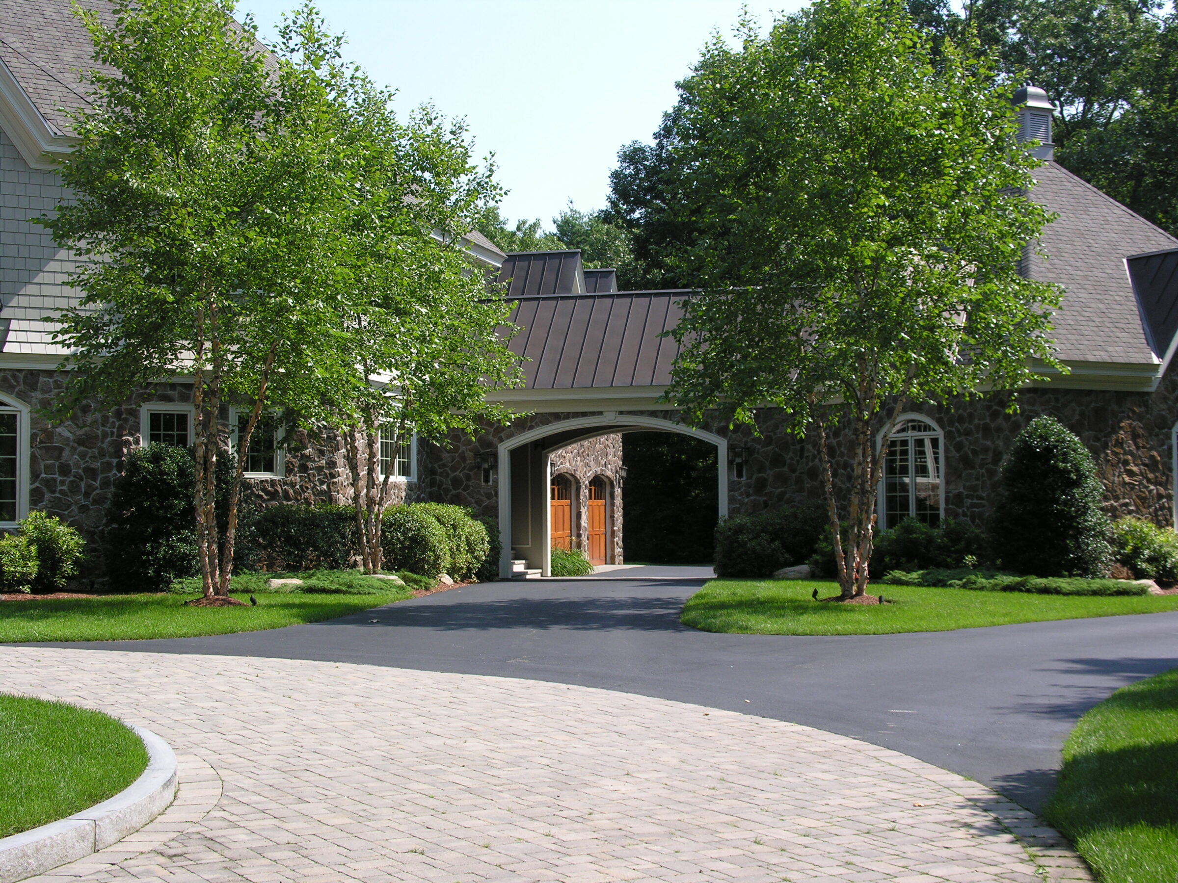 A stone house with an archway entrance, surrounded by trees and a curved driveway, set in a lush, green landscape.
