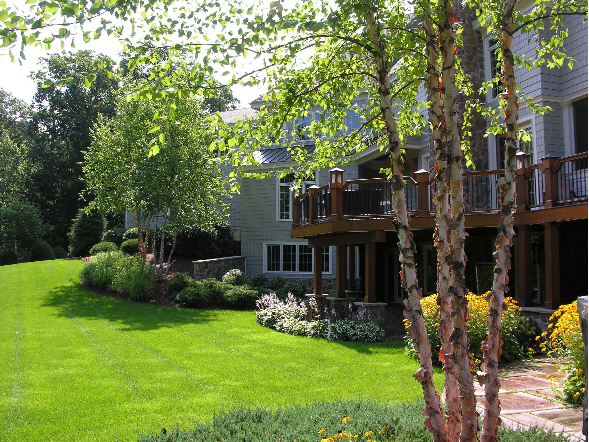 A large house with a well-maintained lawn and garden surrounded by lush trees, featuring a wooden deck and flowering shrubs.