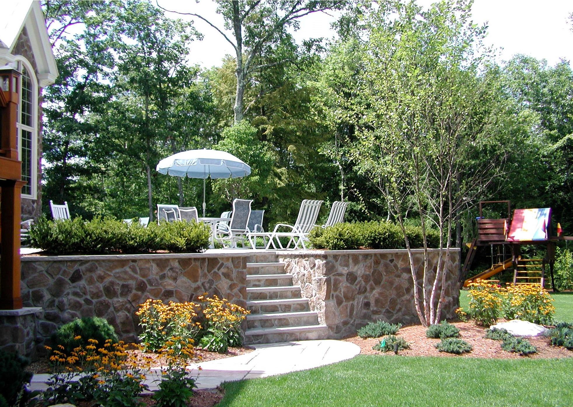 Stone patio with white chairs, umbrella, and garden surrounds. Steps lead to grassy area featuring a child's play structure, bordered by trees.