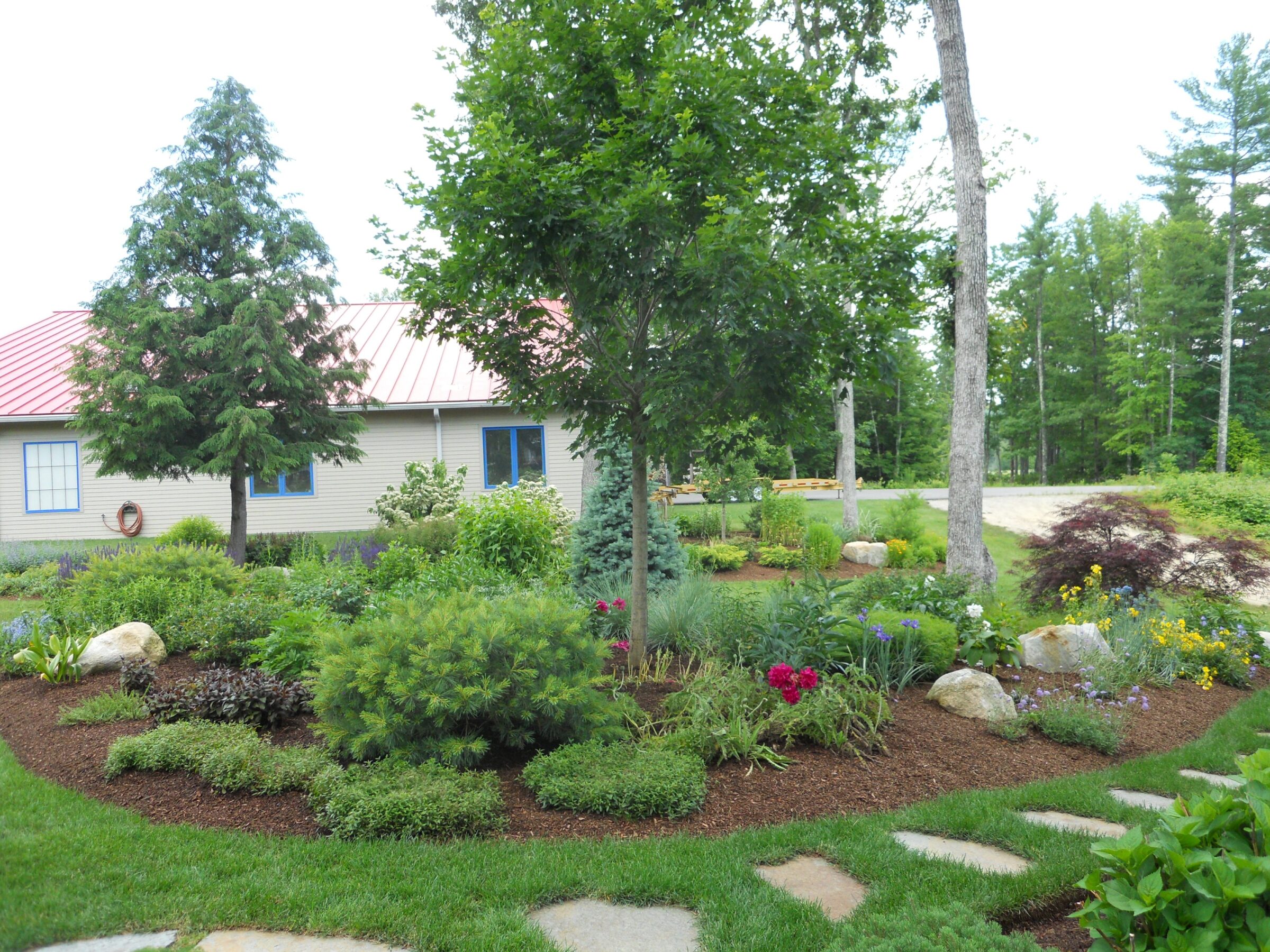 A lush garden features diverse plants and flowers with stepping stones, surrounded by trees. A barn-like structure stands in the background.