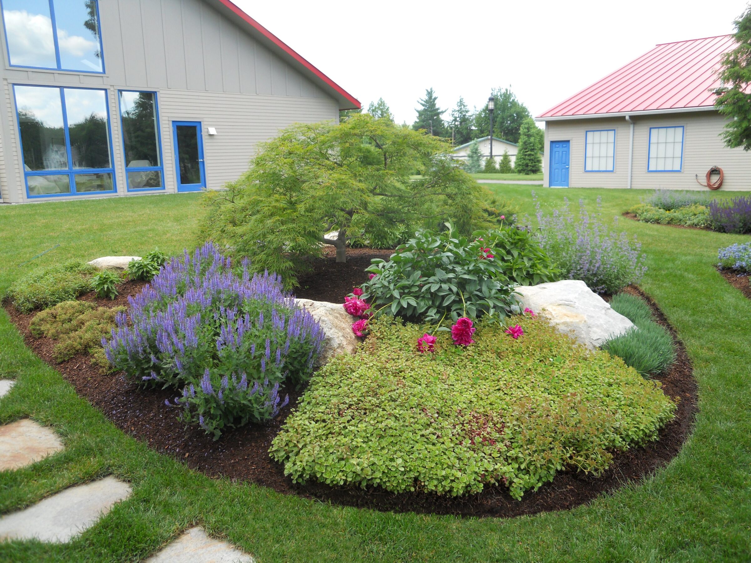 A landscaped garden with colorful flowers and shrubs surrounds two modern buildings with red roofs, blue doors, and large windows.