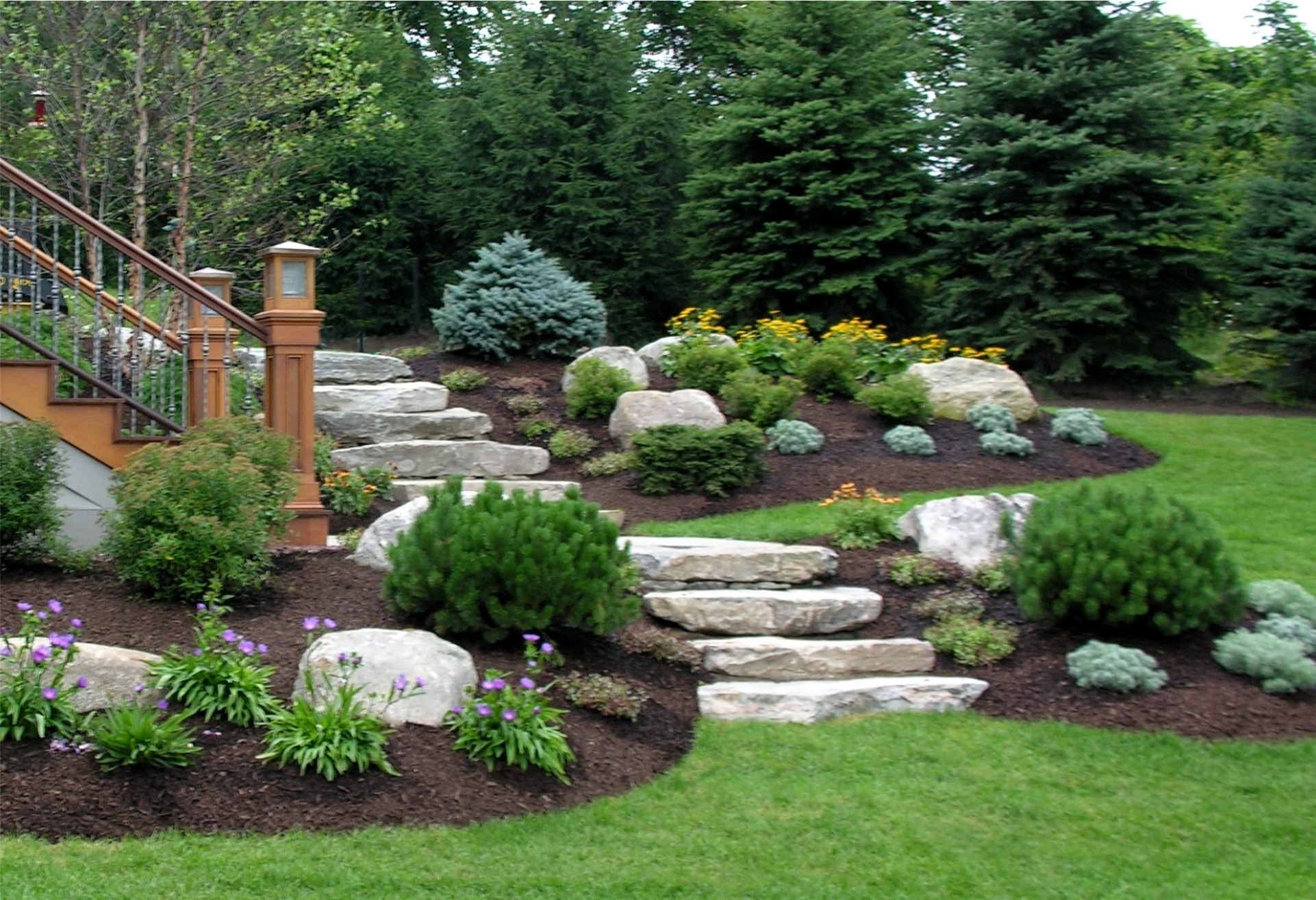 A beautifully landscaped garden features stone steps, vibrant flowers, and manicured shrubs. Wooden railing is visible on the left, complementing the greenery.