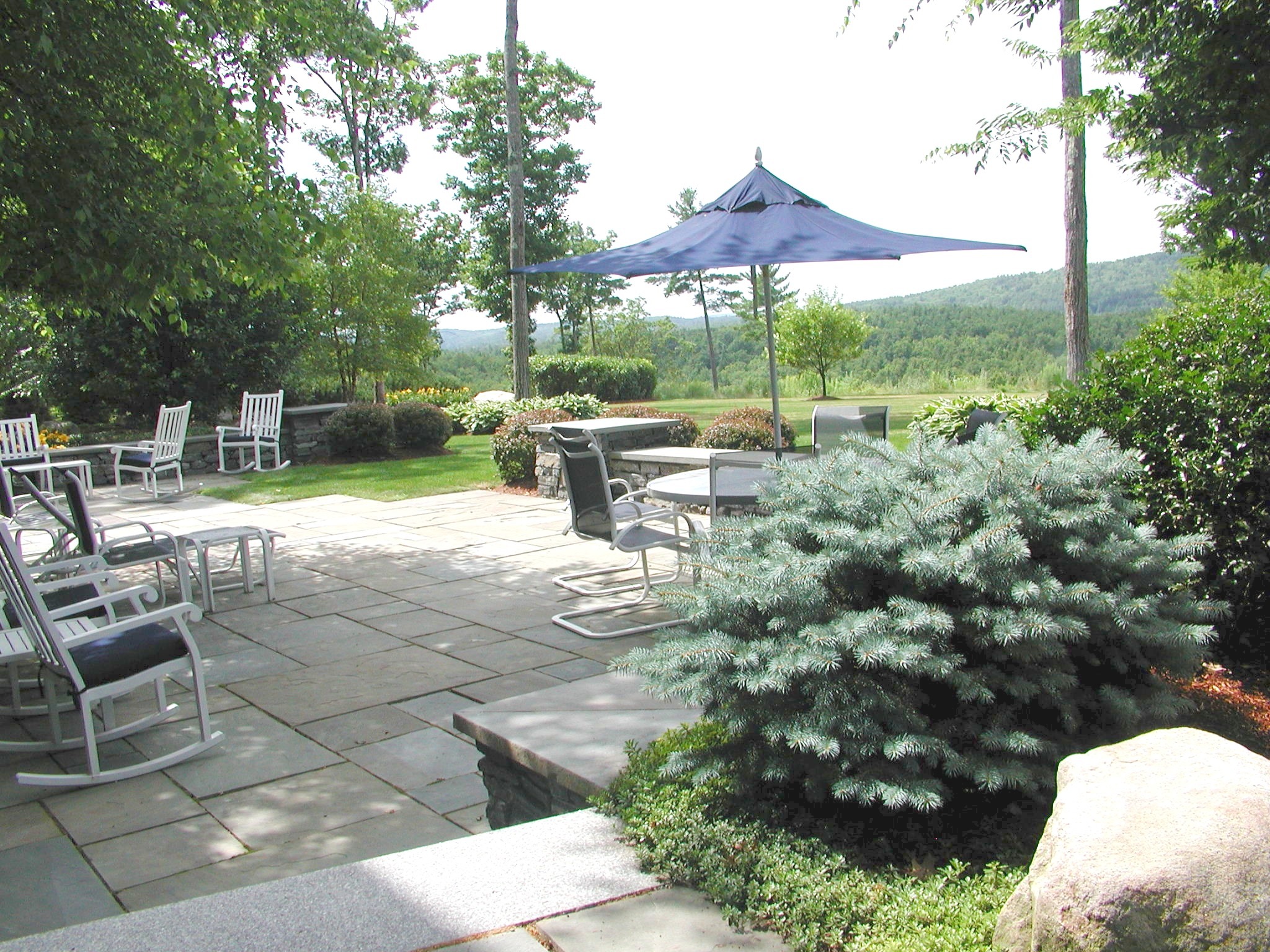 A tranquil patio with chairs, tables, and an umbrella overlooks lush greenery and hills, surrounded by trees and neat shrubbery.