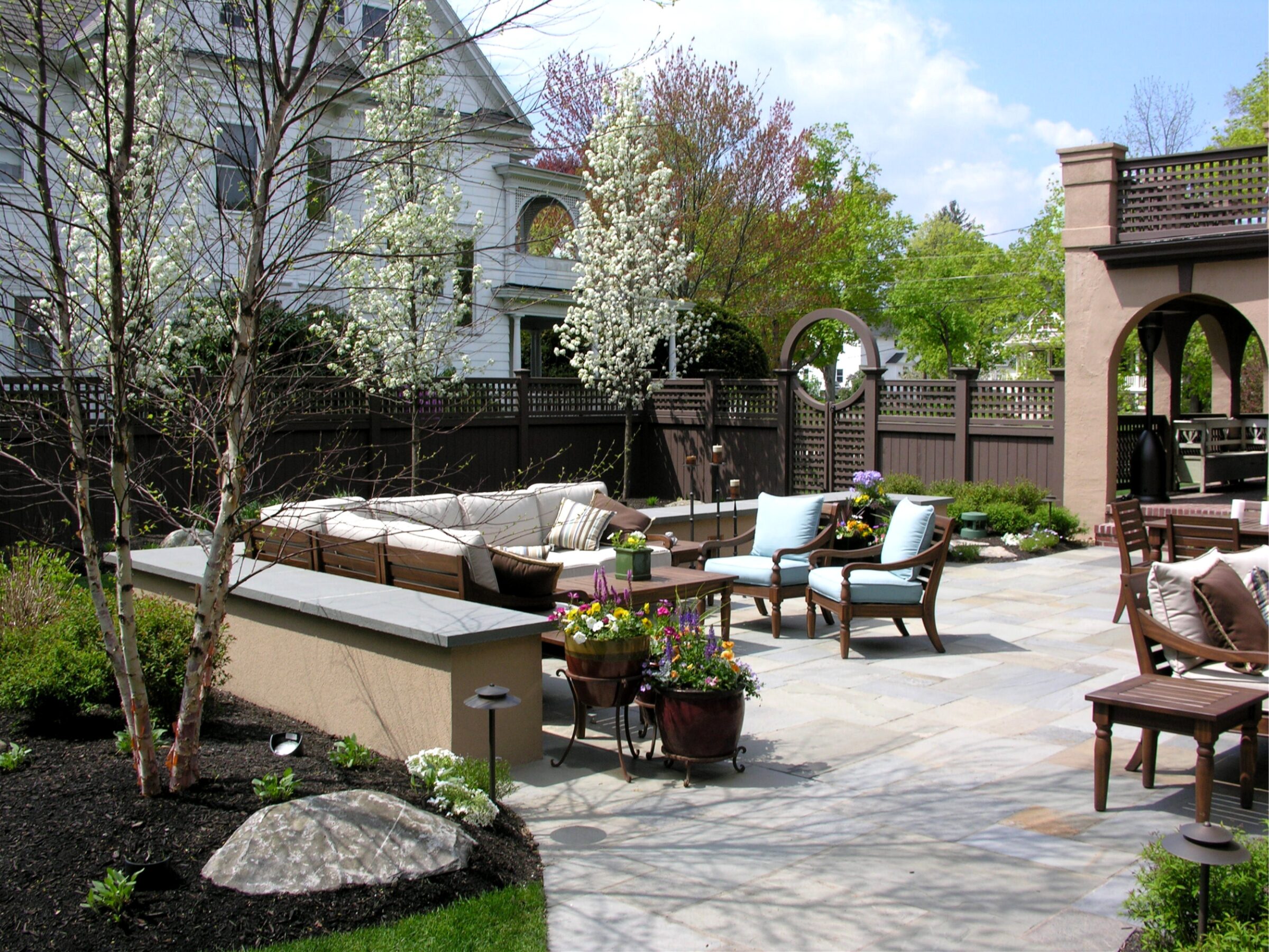 Charming backyard with patio seating, blooming trees, and a variety of flowers. Spacious outdoor area surrounded by a decorative fence and archways.
