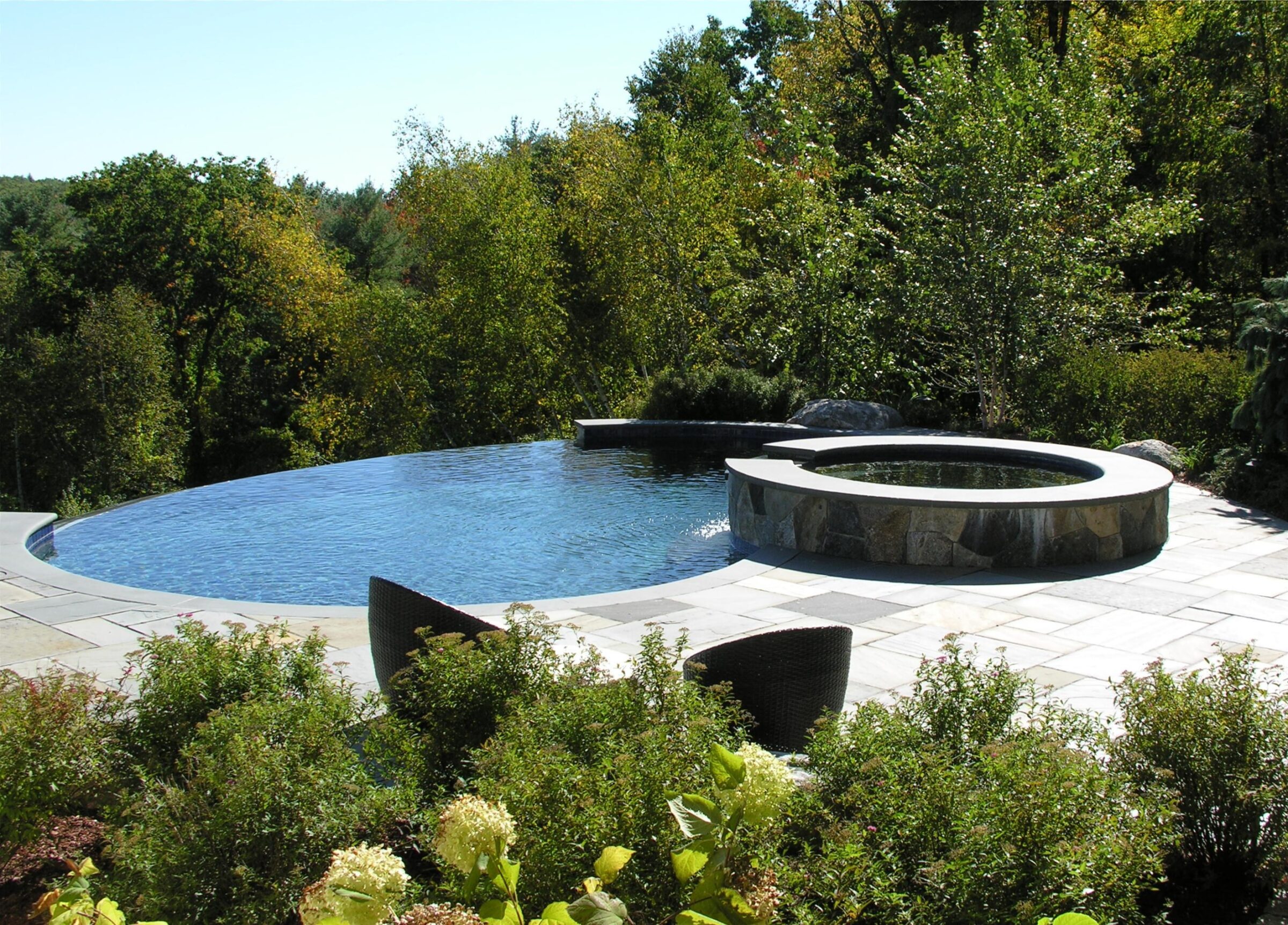 An infinity pool and hot tub overlook lush greenery, surrounded by stone tiles and garden foliage, under a clear sky.