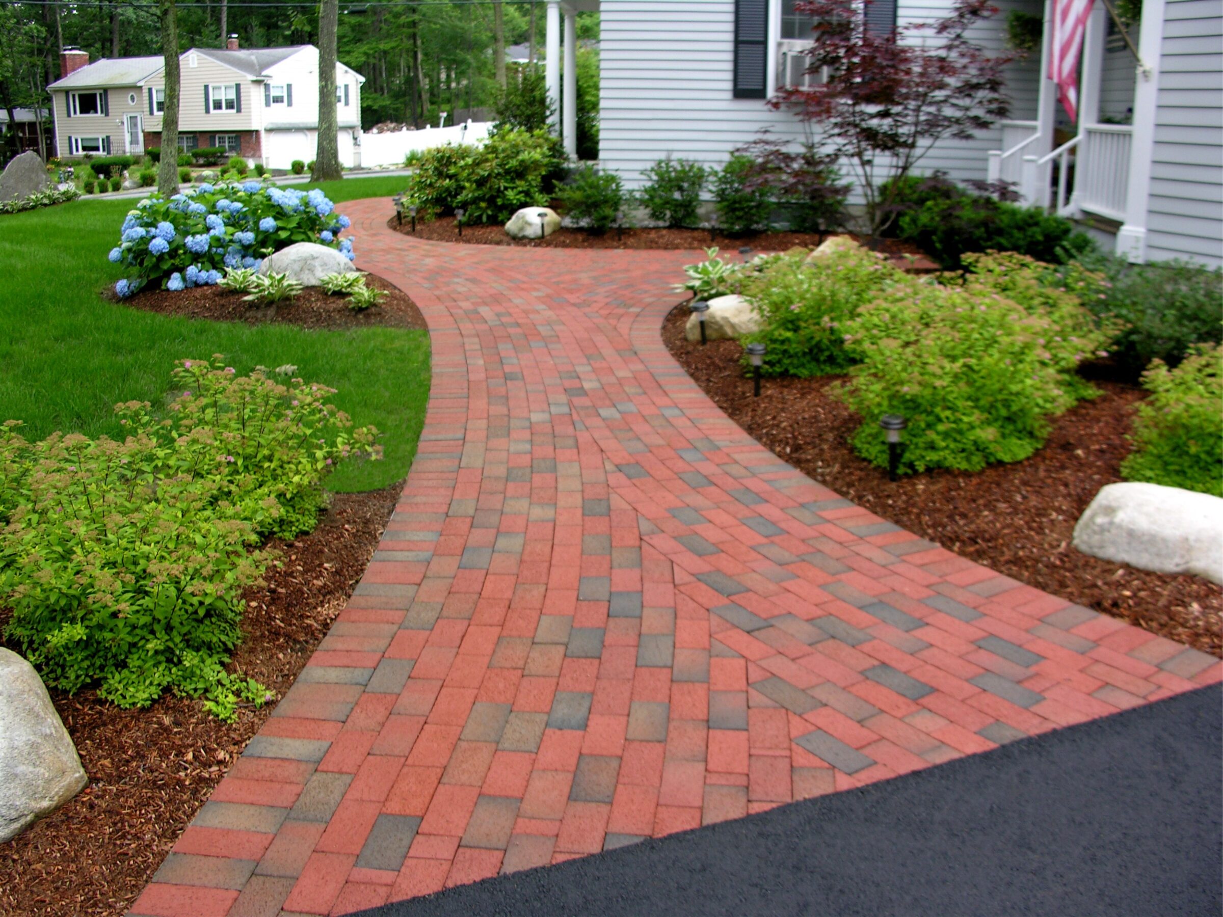 A neatly landscaped front yard features a red brick walkway, green shrubs, and hydrangeas, leading to a house with gray siding.