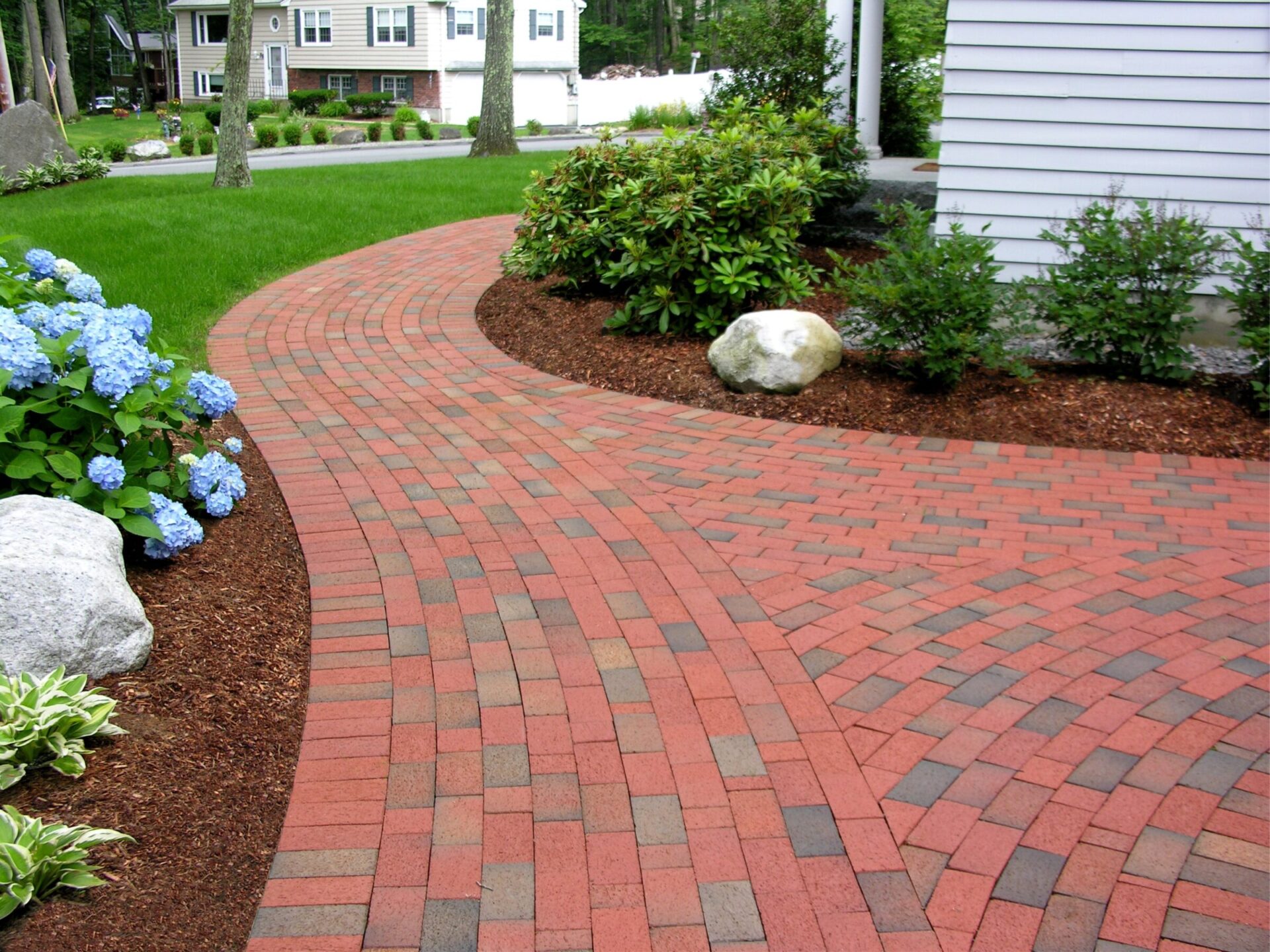 A brick pathway curves through a landscaped garden with shrubs and flowers, leading to a white house. Trees and a grassy yard surround it.