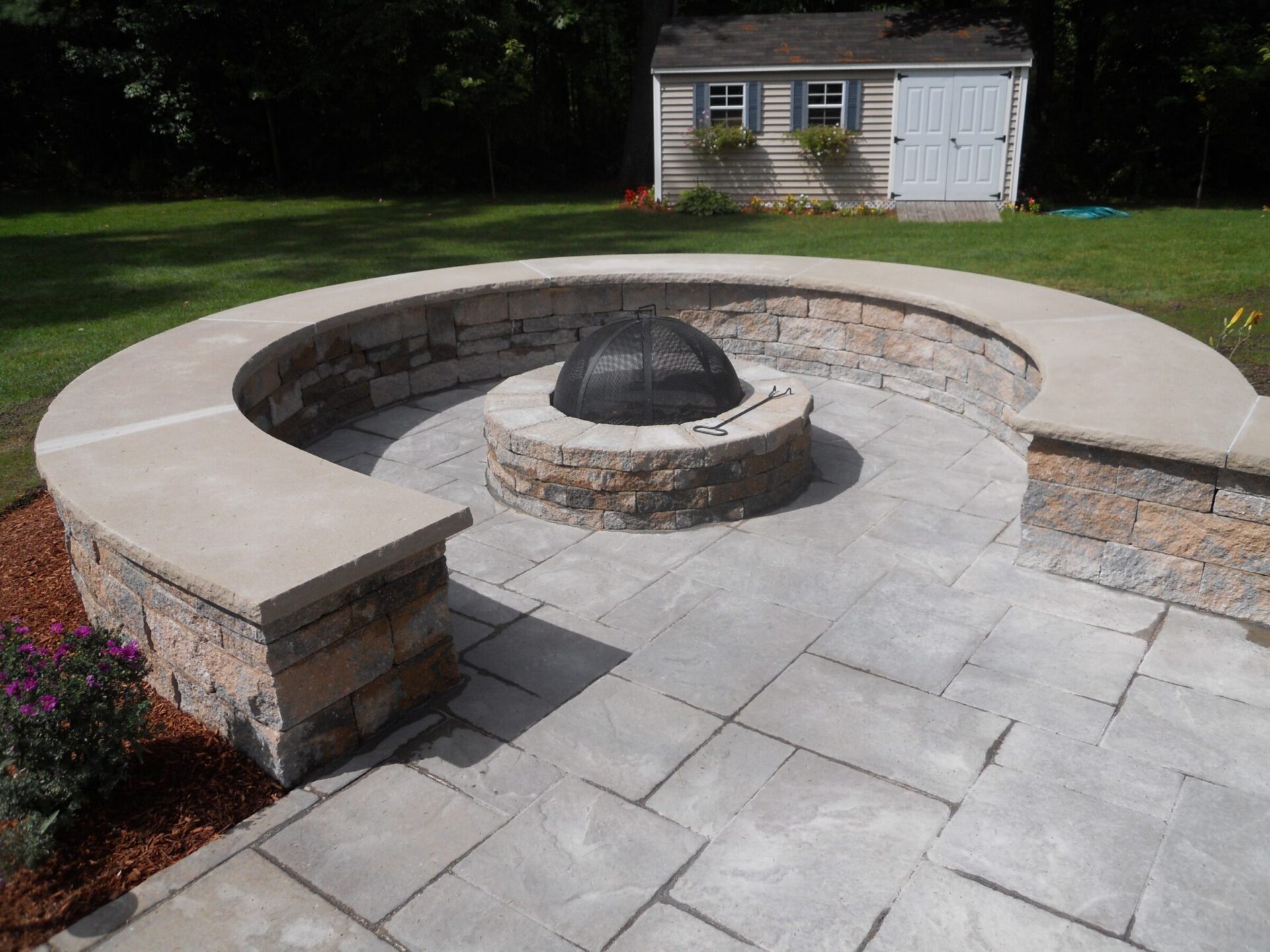 Circular stone fire pit with seating area on a paved patio. Small shed and green lawn in the background under clear skies.