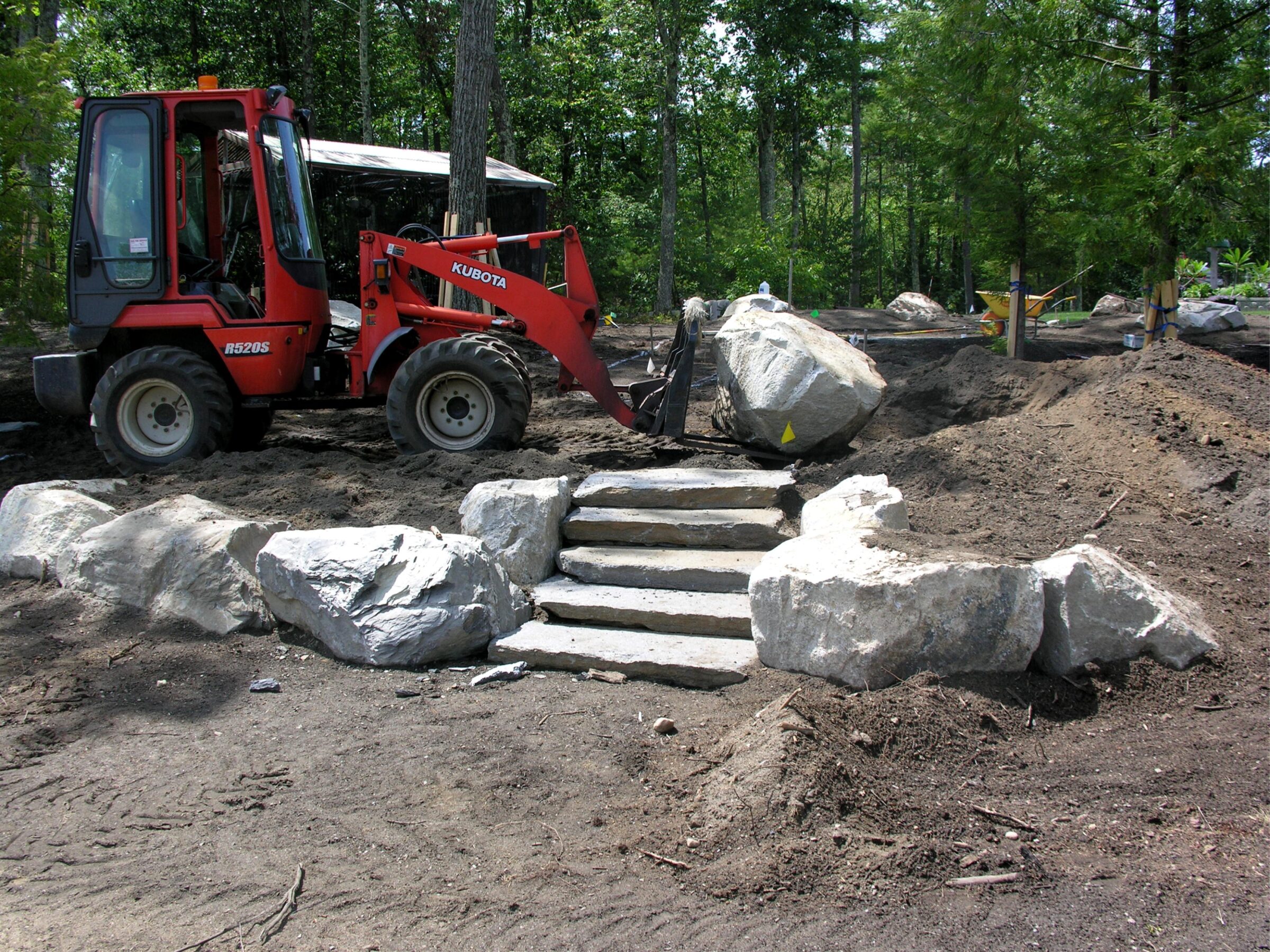 A red tractor positions large rocks near newly constructed stone steps in a forest setting, with trees and dirt surrounding the area.