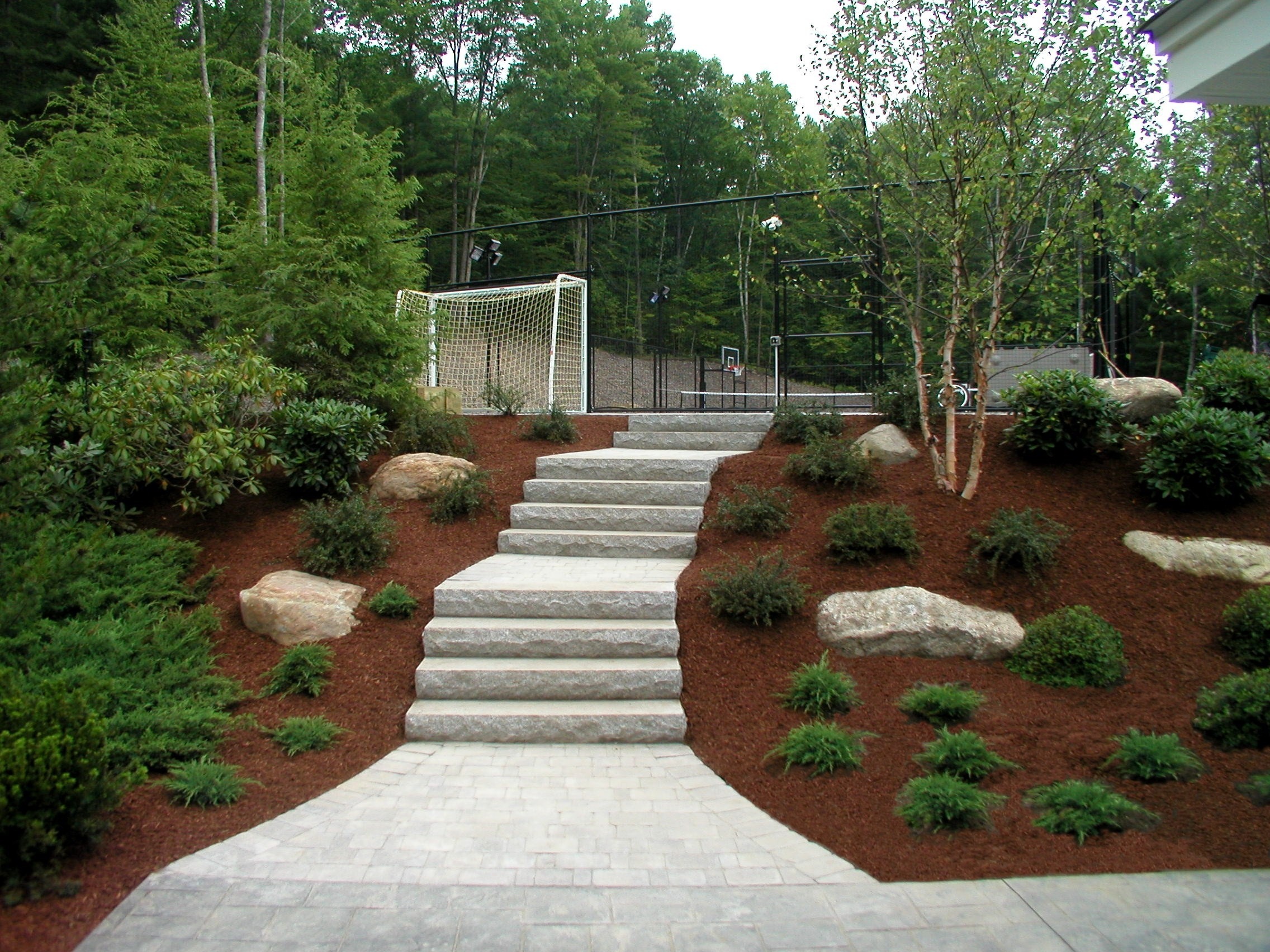Stone steps lead through a landscaped garden with shrubs and trees, towards a soccer field with a goal, surrounded by lush greenery.