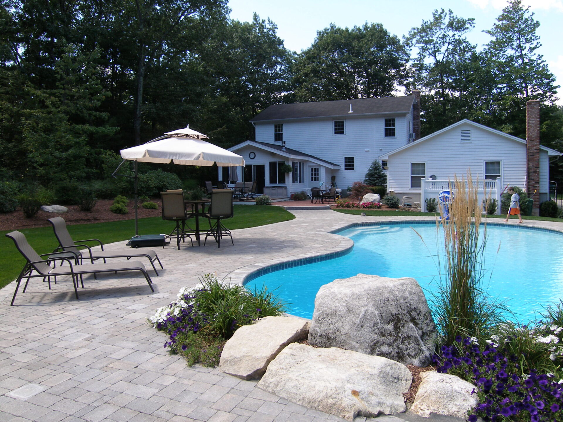 A backyard scene with a pool, lounge chairs, umbrella, and a white house surrounded by trees and landscaped gardens. No people visible.