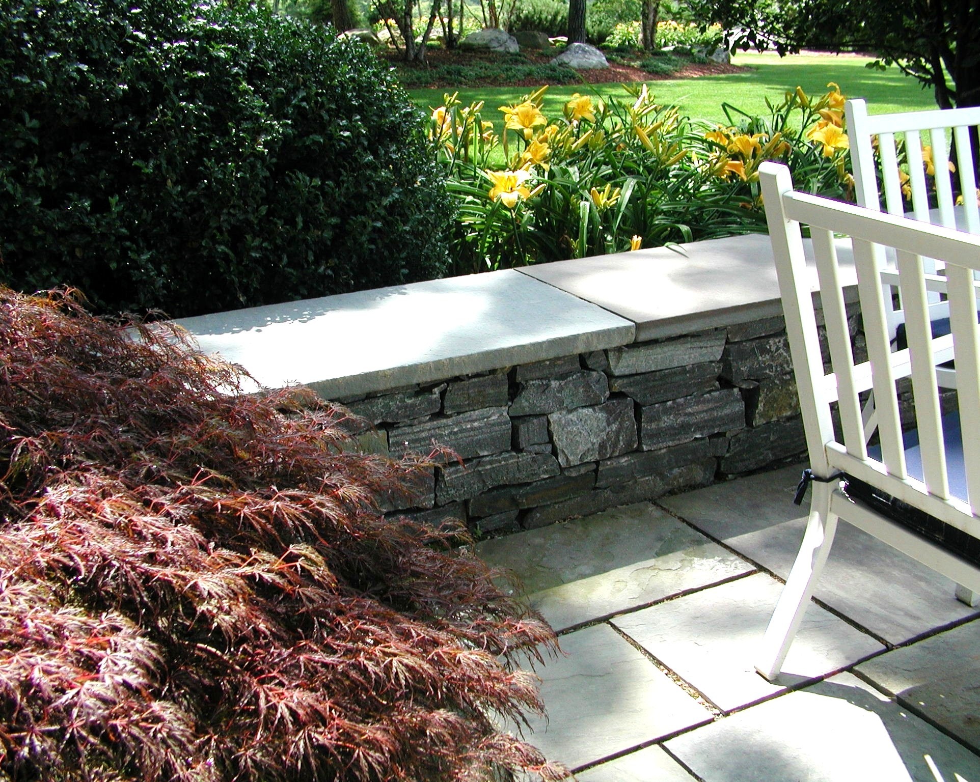 A sunlit garden features a white wooden chair, stone wall, vibrant yellow flowers, and lush greenery, creating a peaceful outdoor setting.