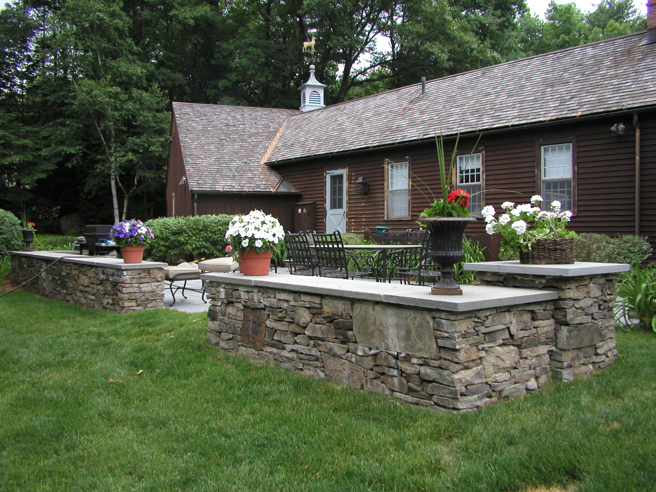 Stone patio with flower pots and outdoor furniture beside a rustic wooden house. Lush trees and grass surround the serene setting.