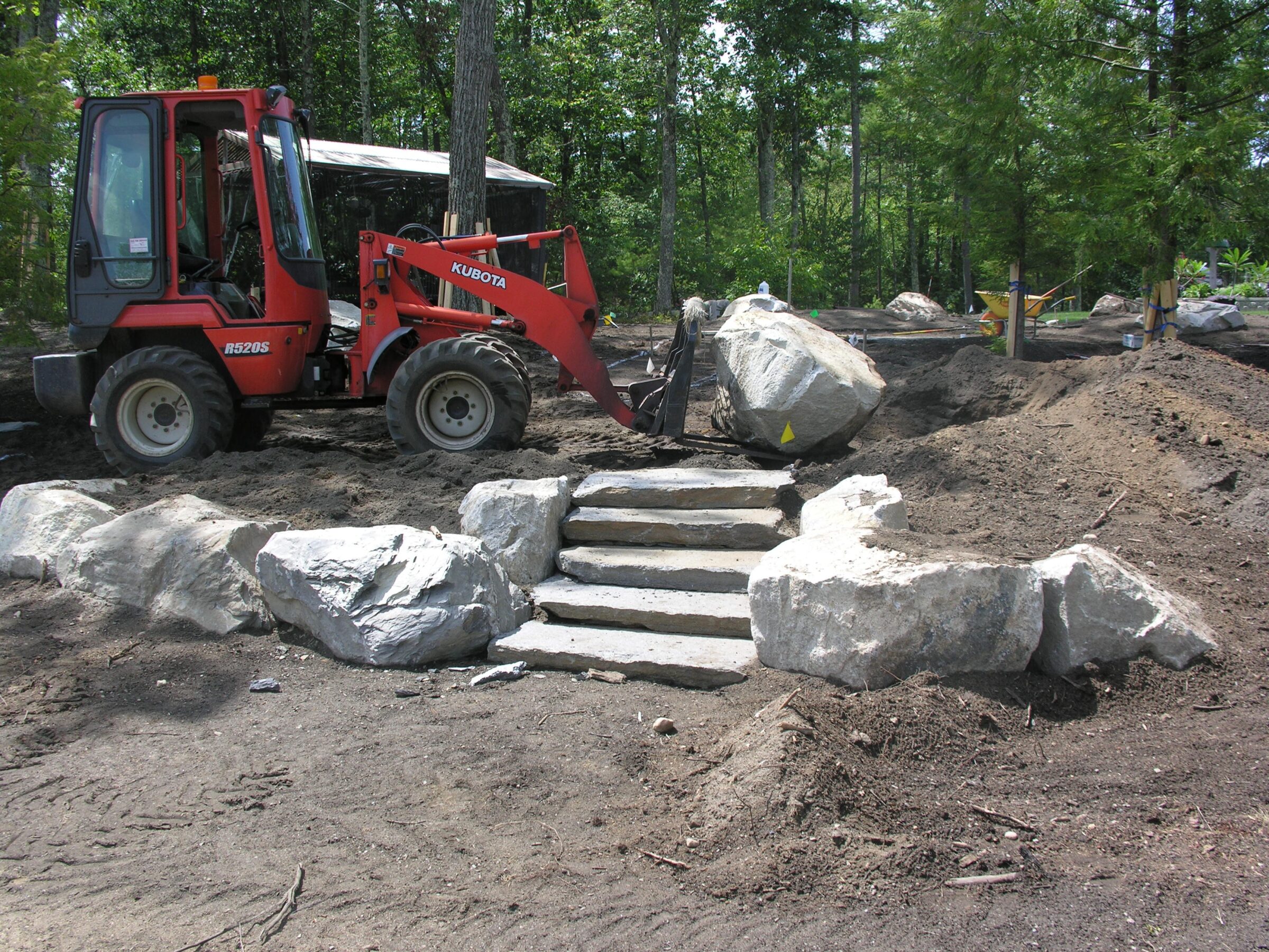 A red Kubota forklift moves a large boulder near newly constructed stone steps in a wooded area with greenery and exposed earth.