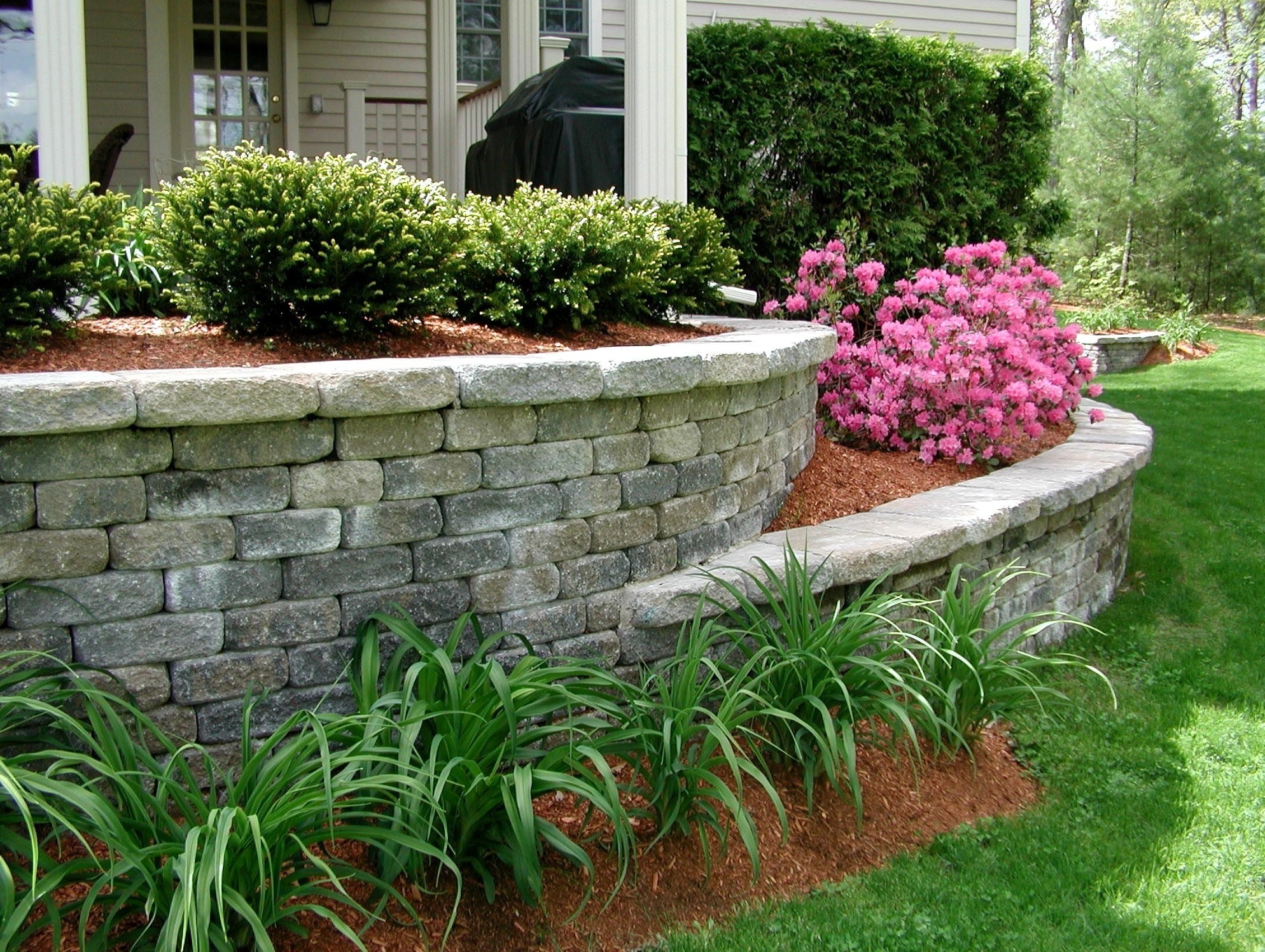A garden with a stone retaining wall, pink flowers, and green plants surrounds a white house porch, creating a charming landscape.