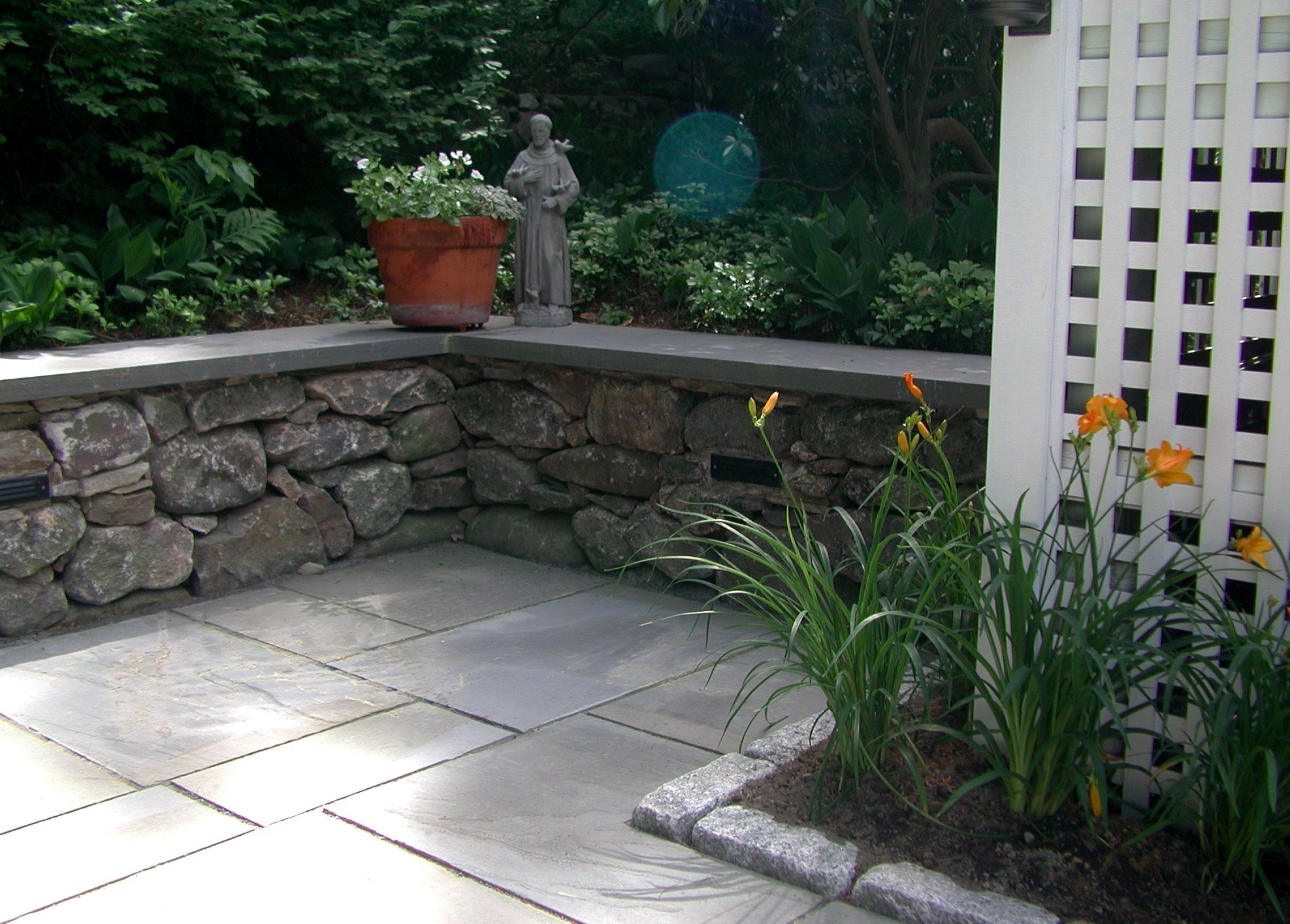 A tranquil garden nook with a stone bench, statue, potted flowers, and vibrant lilies beside a white trellis. Peaceful and inviting atmosphere.