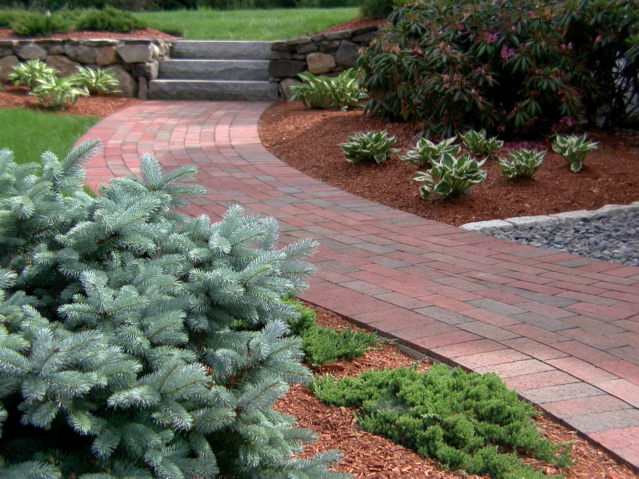 A curved brick pathway winds through a landscaped garden with greenery, shrubs, and stone borders, leading to stone steps amidst mulch beds.