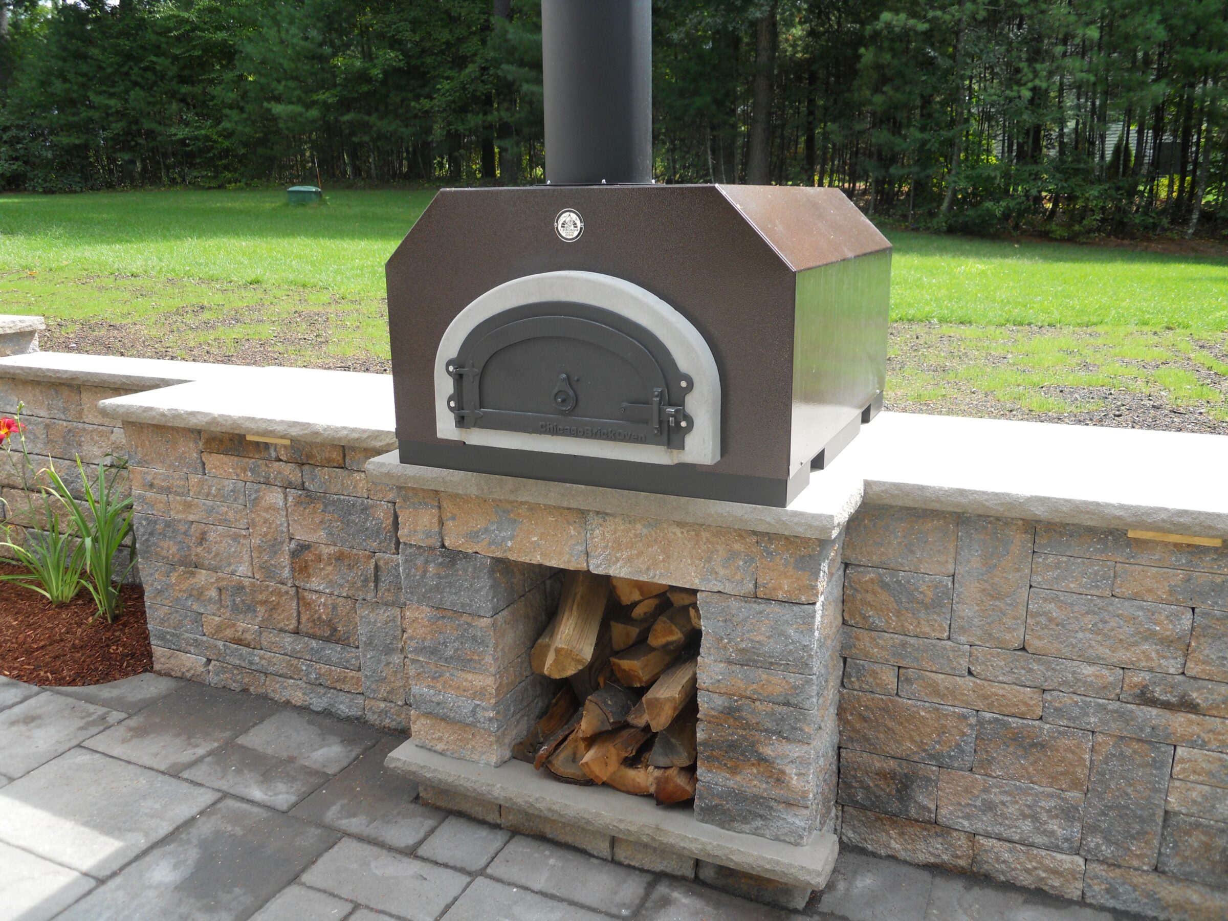 A brick outdoor pizza oven stands on a stone counter, with firewood stored beneath. Surrounded by paved patio and lush greenery.