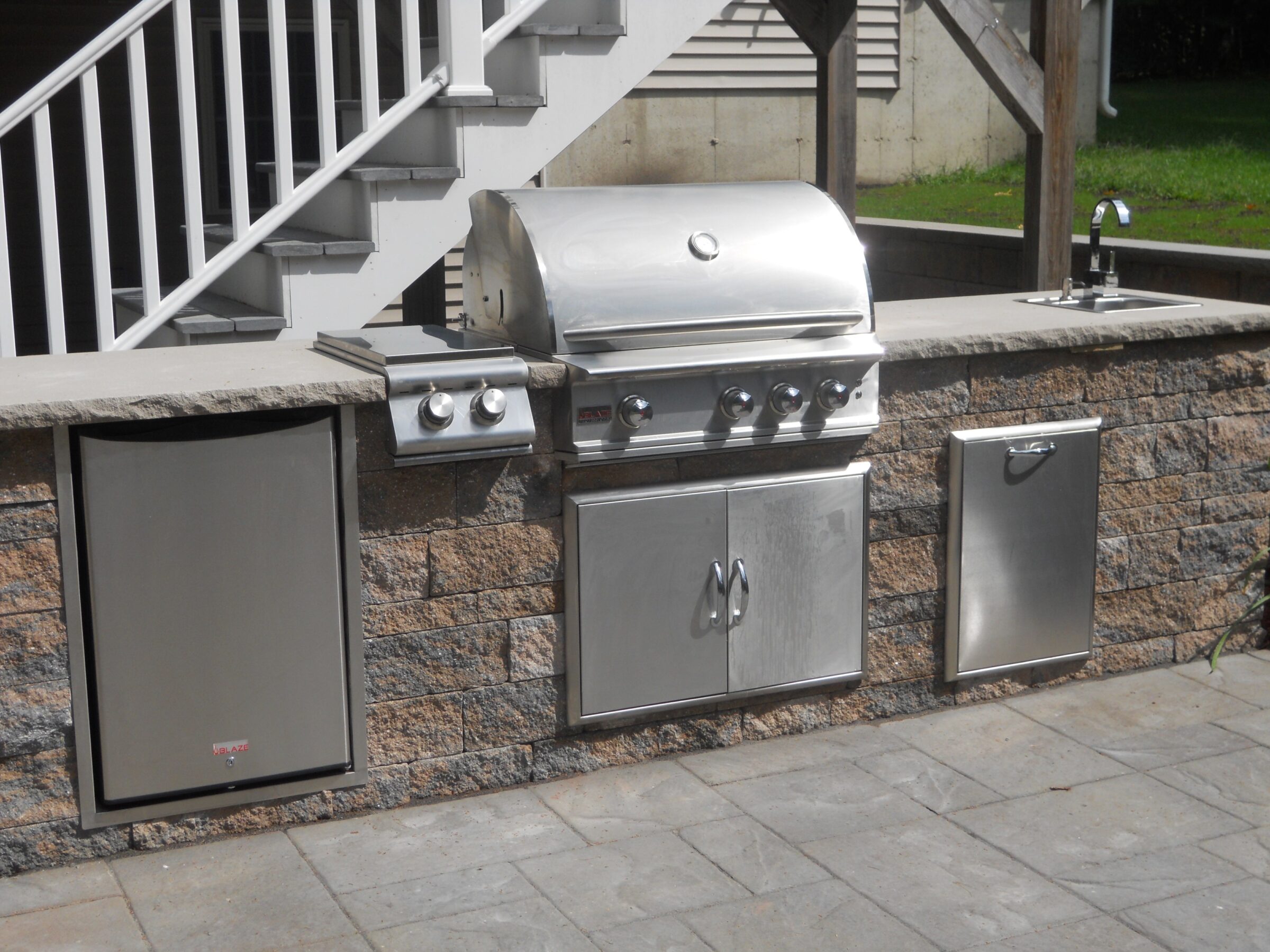 An outdoor kitchen setup featuring a stainless steel grill, fridge, and sink, placed on a stone counter with a staircase behind.