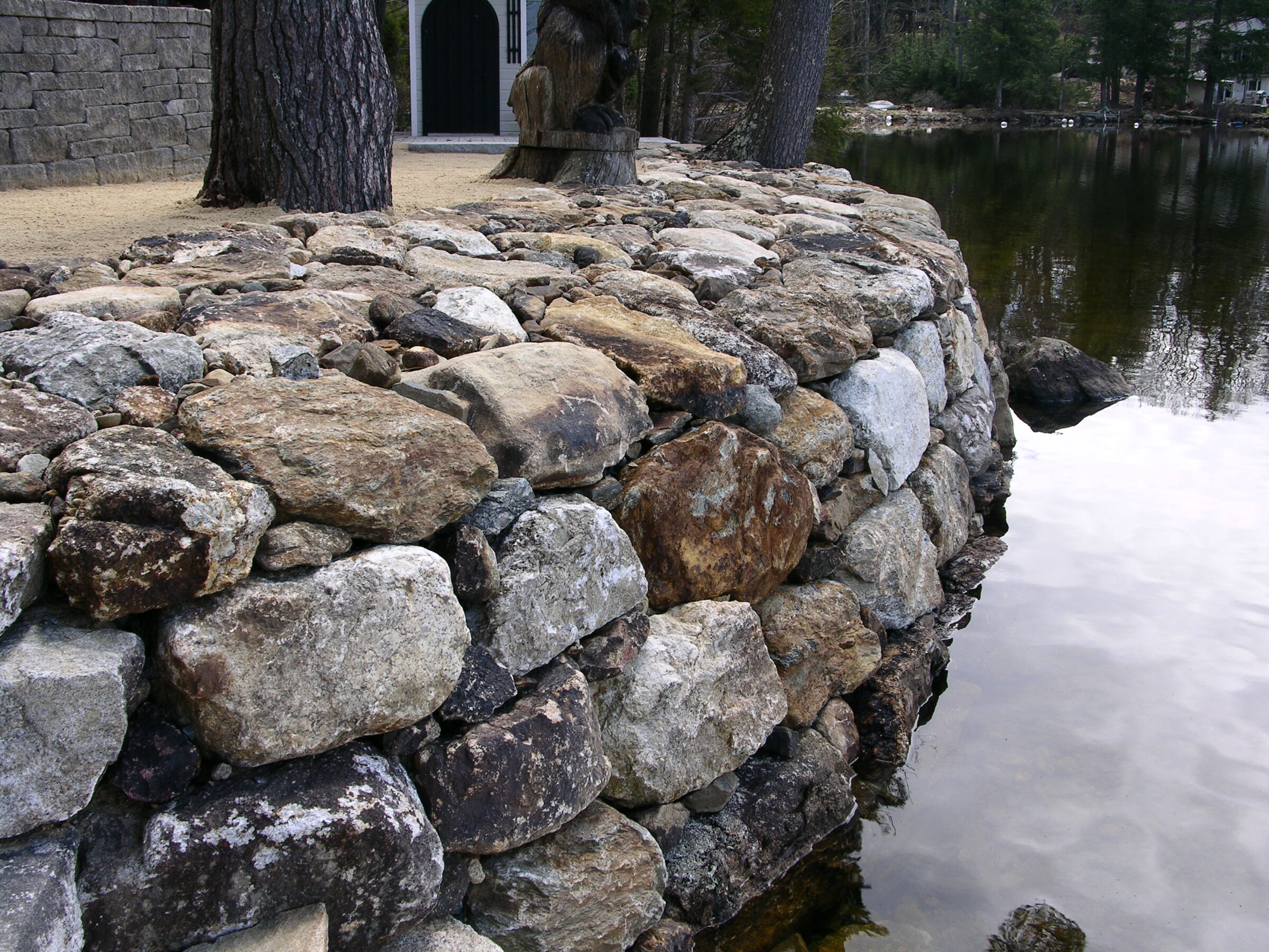 A stone wall curves alongside a calm shoreline, with trees and a partial view of a structure in the background.