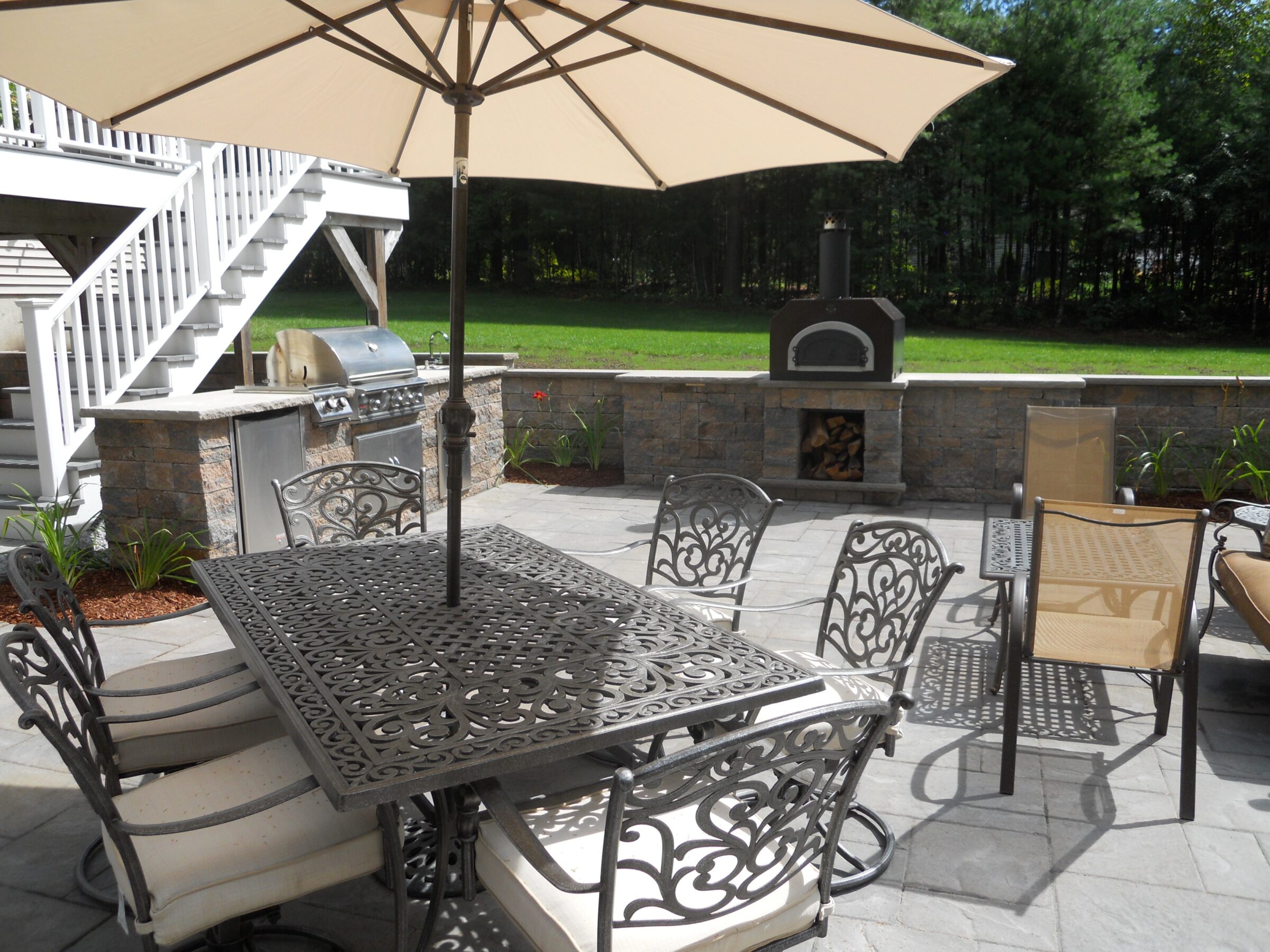A patio with ornate metal furniture, large umbrella, outdoor grill, and fireplace set against a backdrop of lush green trees and grass.