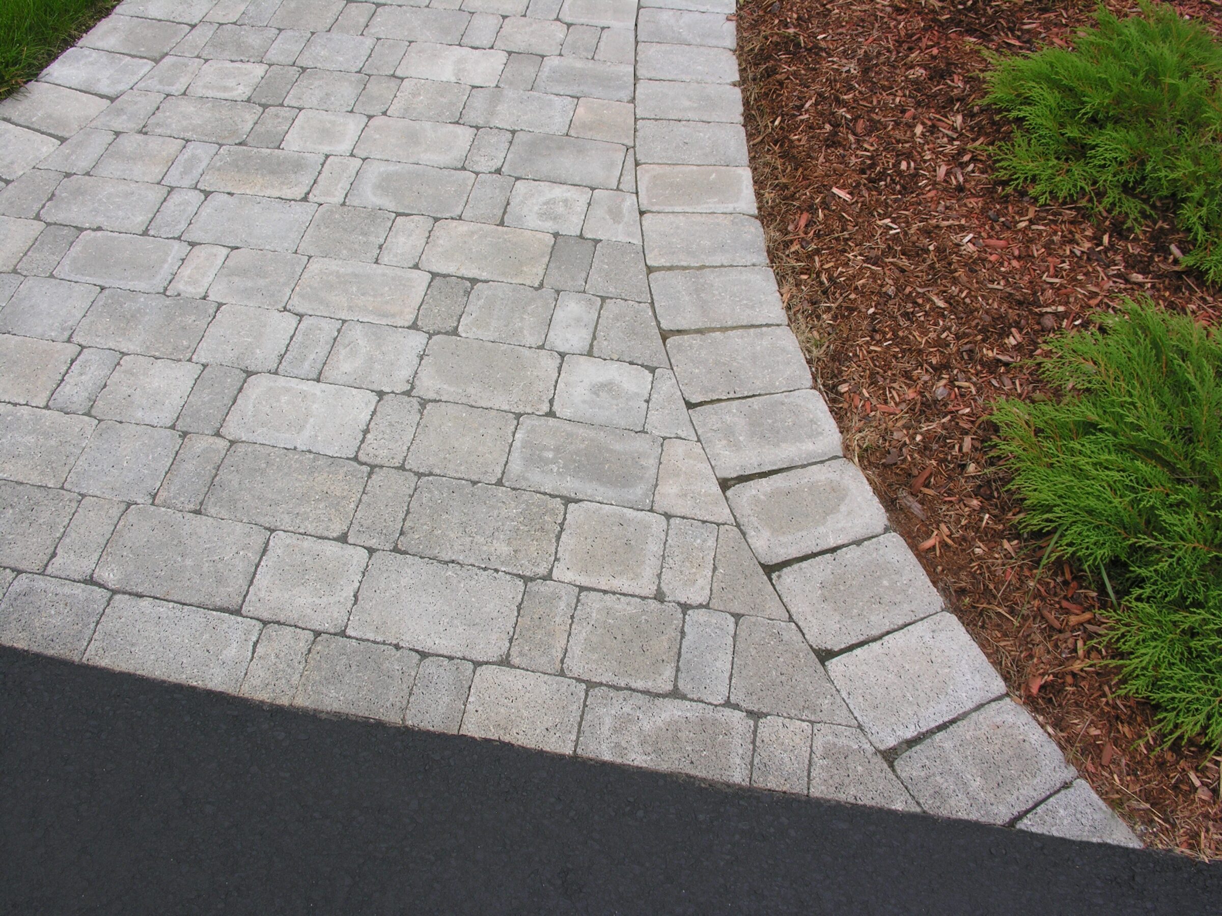 Curved stone pathway with neat grey bricks, bordered by mulch and green shrubs, adjacent to a strip of blacktop pavement.