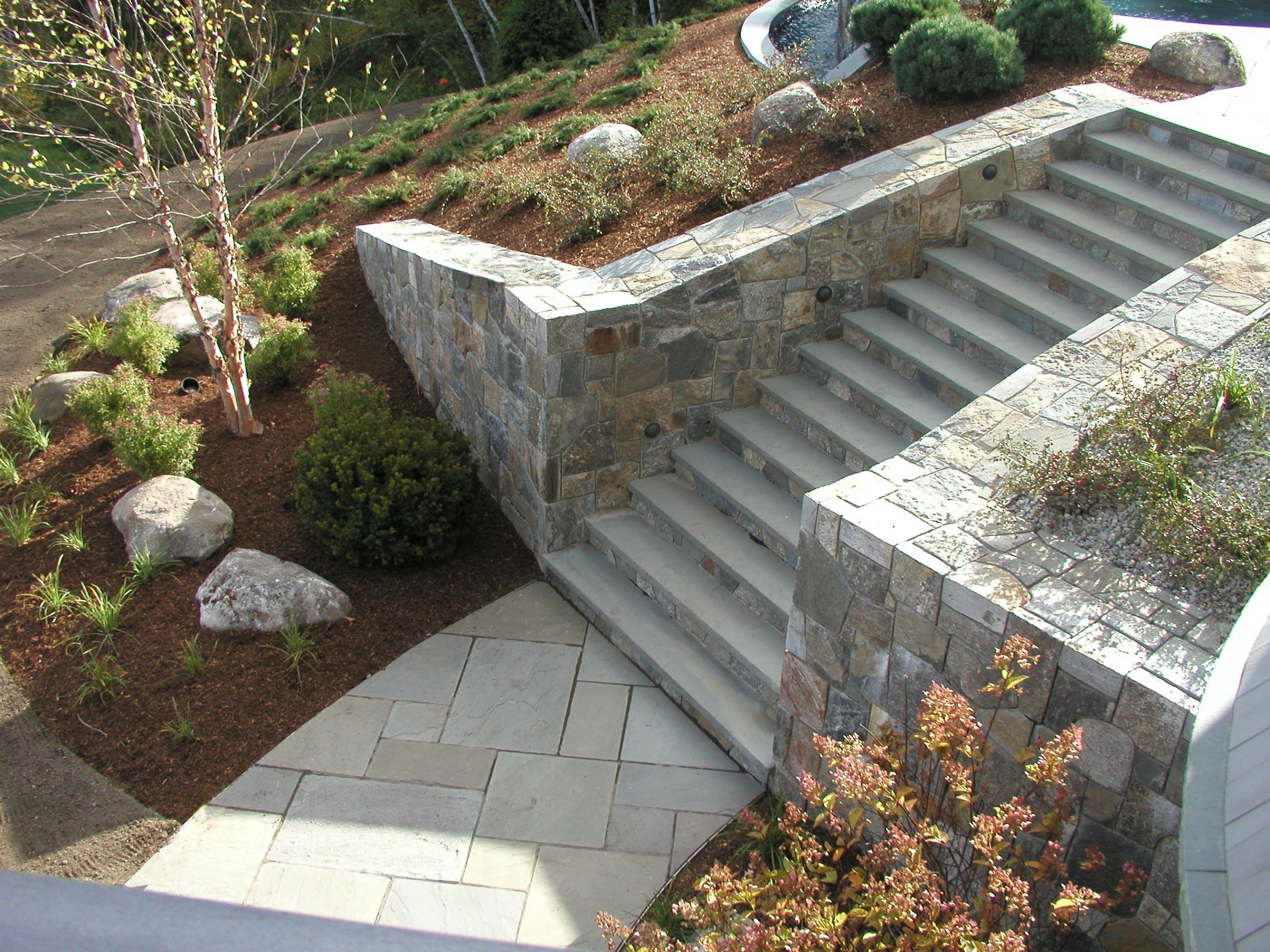 Stone stairs with surrounding landscaped garden, featuring shrubs and small trees. The area includes a curved stone wall and tiled pathway.