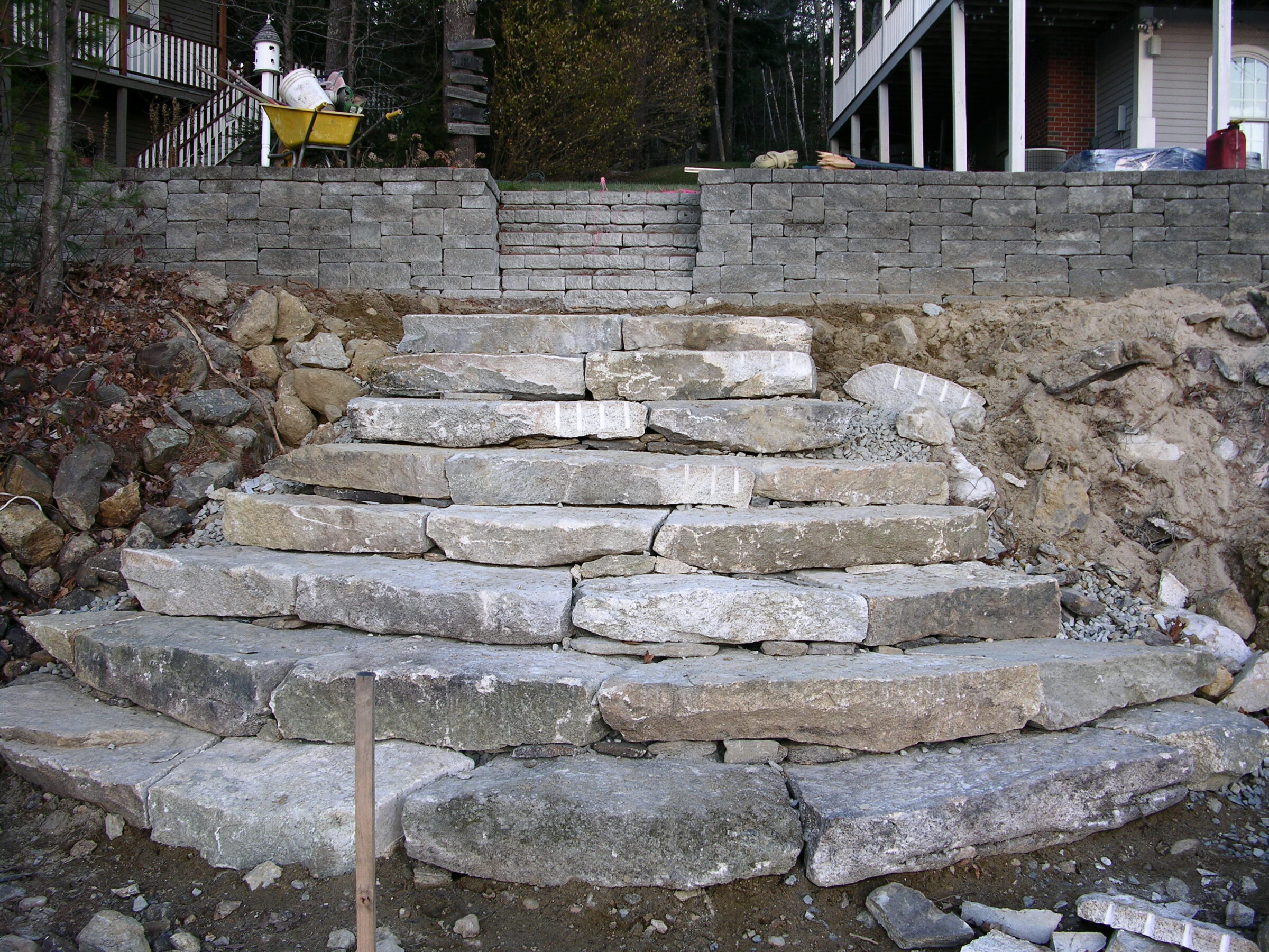 Stone steps under construction, built into a hillside with stone retaining walls. Nearby, a wheelbarrow and trees are visible in the background.