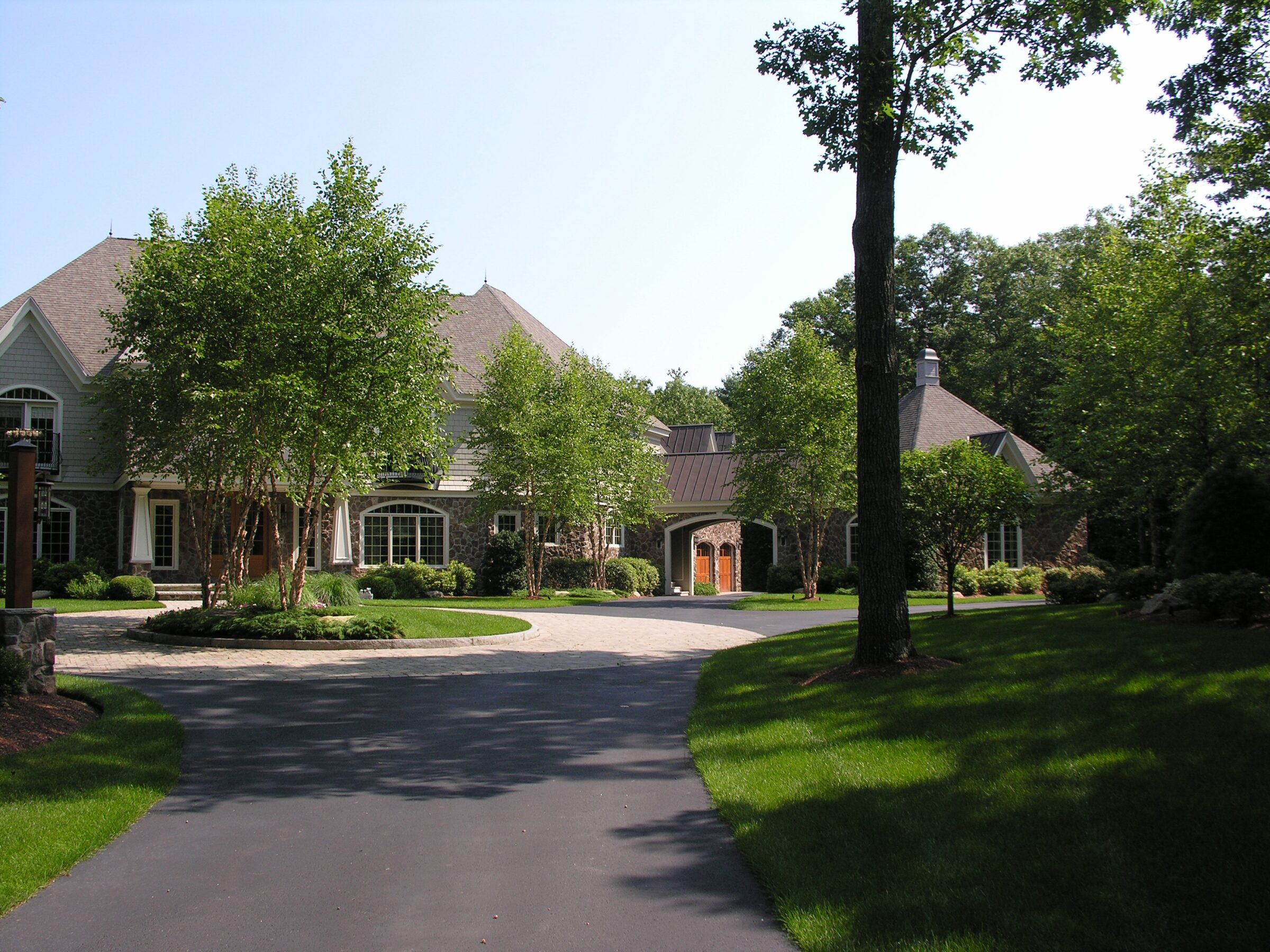 A large suburban house is surrounded by trees and a well-maintained lawn, with a curved driveway leading to the entrance.