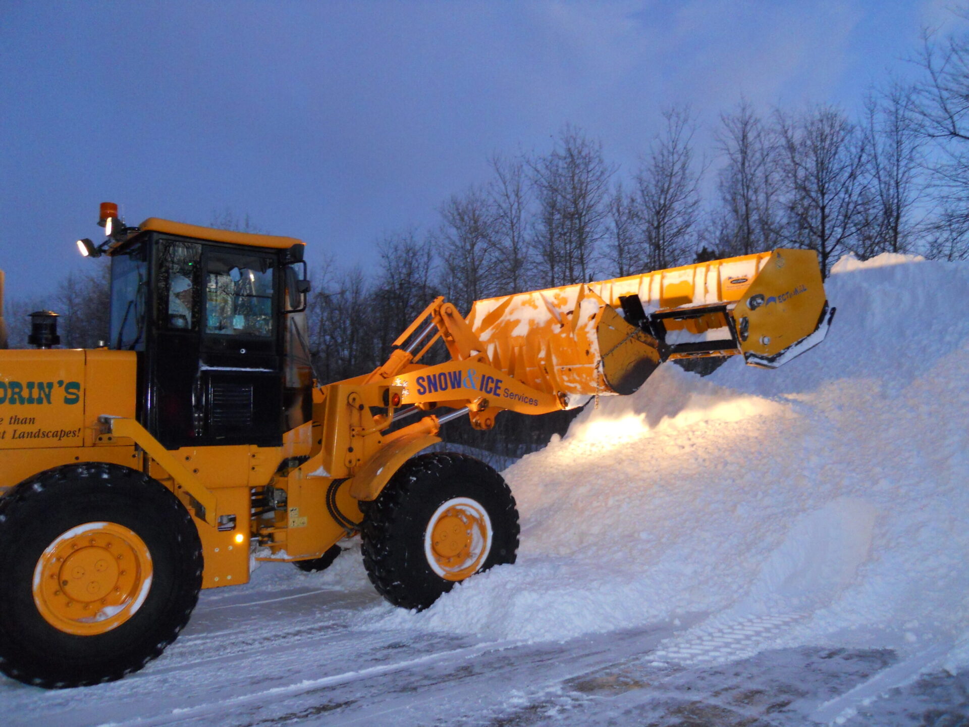 A snow removal vehicle clears a large snowpile at dusk. Trees line the background, and lights from the vehicle illuminate the snow.