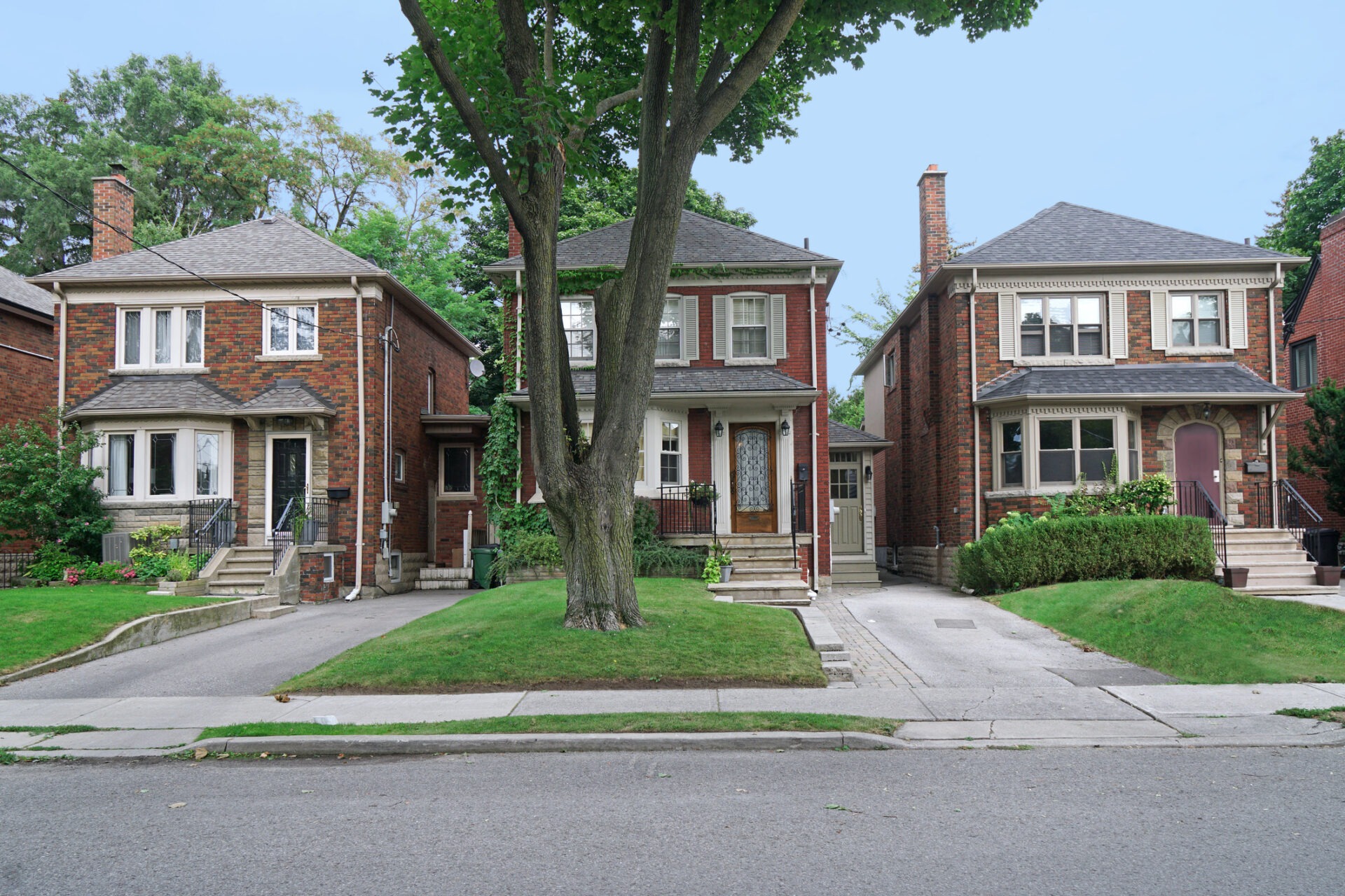 Three brick houses with manicured lawns stand side by side on a residential street. A large tree grows in front of the middle house.