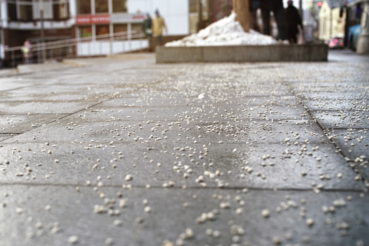 Sidewalk covered with scattered salt grains leading to snow pile, people walking in the background, urban setting, no recognizable landmarks or buildings visible.