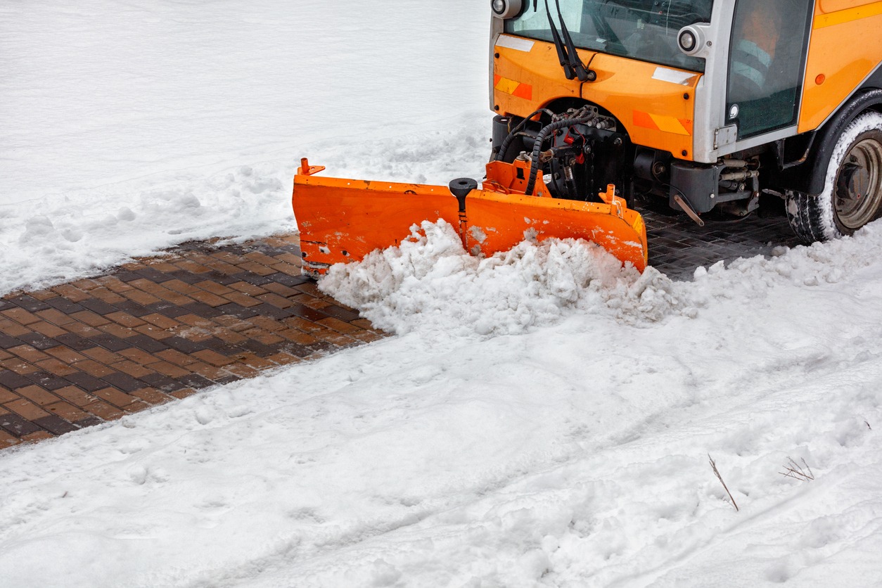 An orange snowplow clearing a brick path surrounded by snow, creating a neat and clean pedestrian walkway in a snowy landscape.