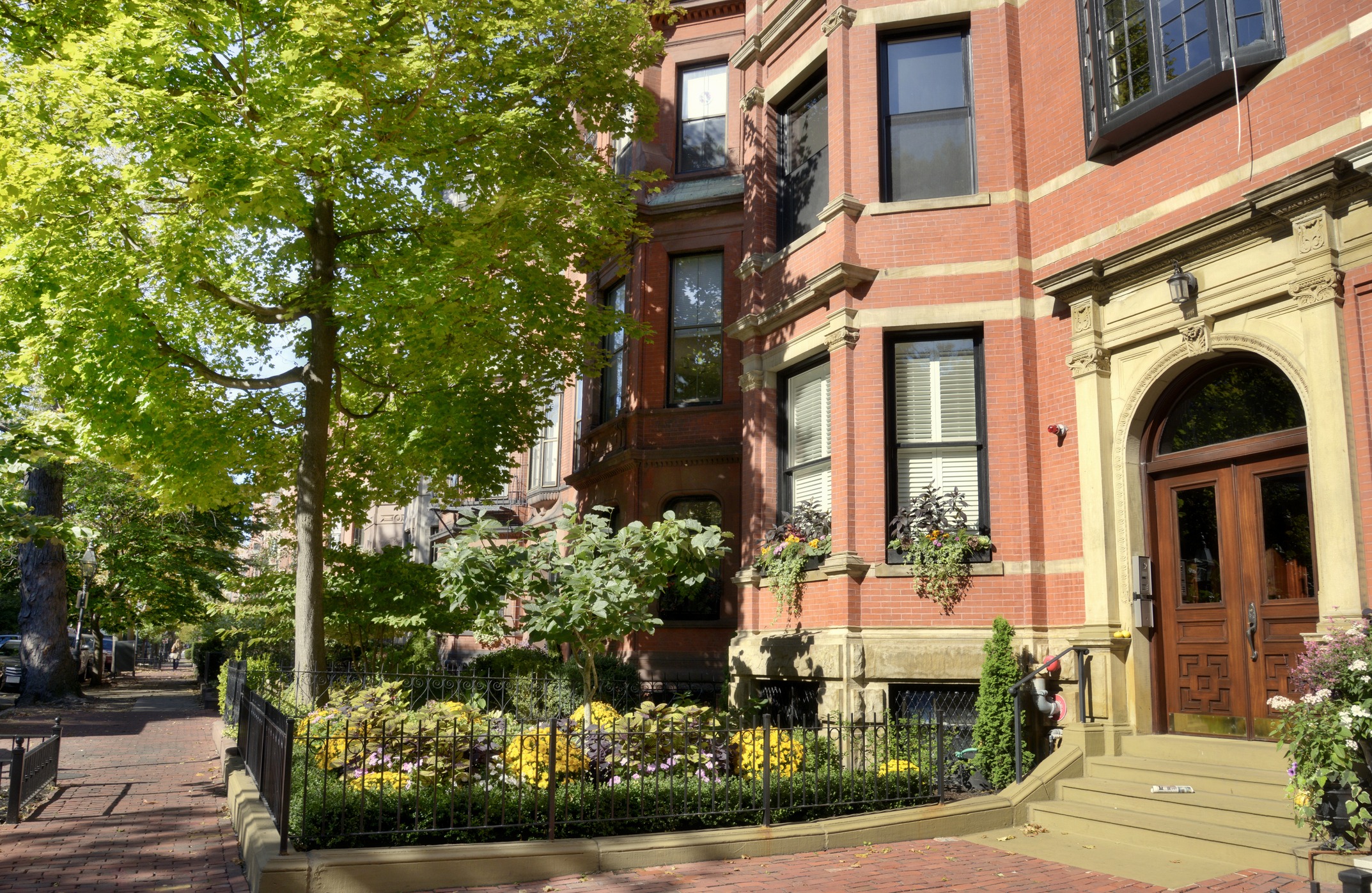 Historic brownstone building with ornate entrance, surrounded by lush greenery and blooming flowers. The brick sidewalk enhances the charming, urban neighborhood ambiance.