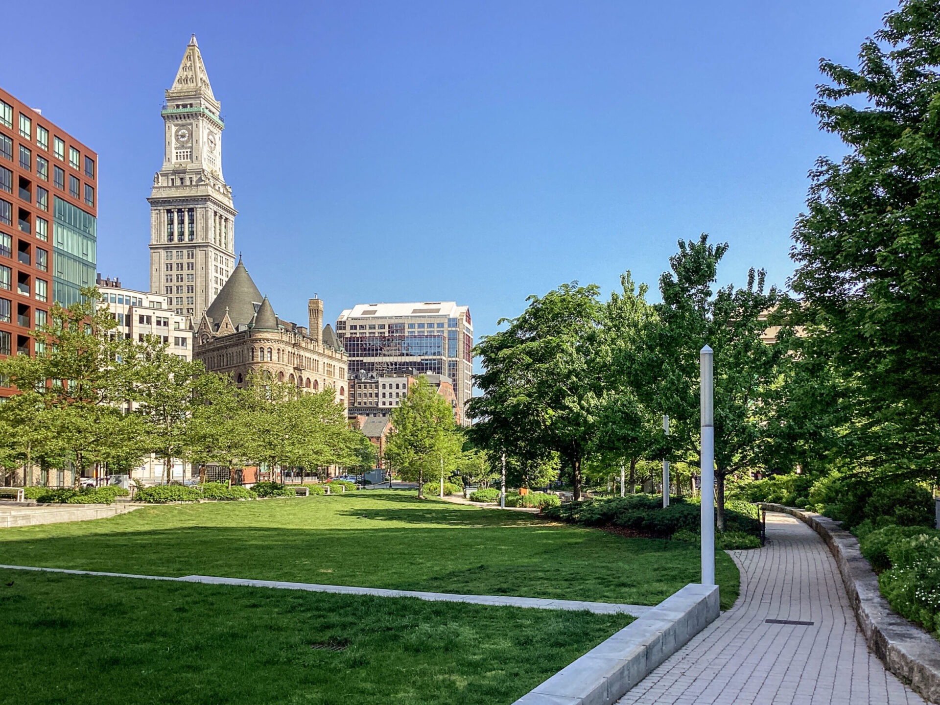 A sunny day at Boston's Christopher Columbus Park, featuring the Custom House Tower, surrounded by greenery and urban buildings, with a walking path.